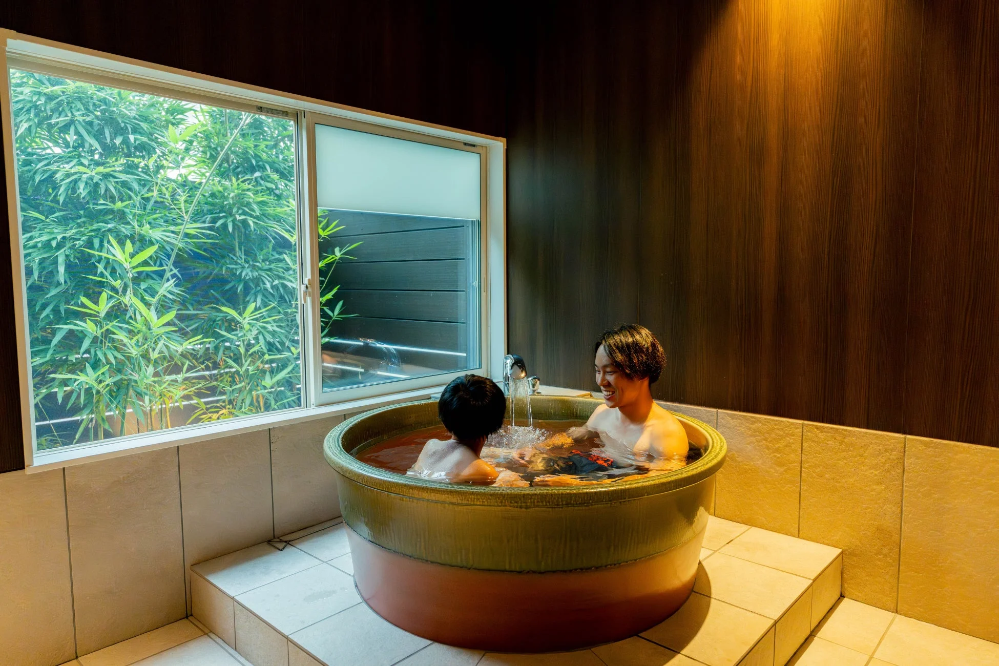 窓の外の竹林を眺めながら、二人の人物が丸い浴槽でくつろいでいる