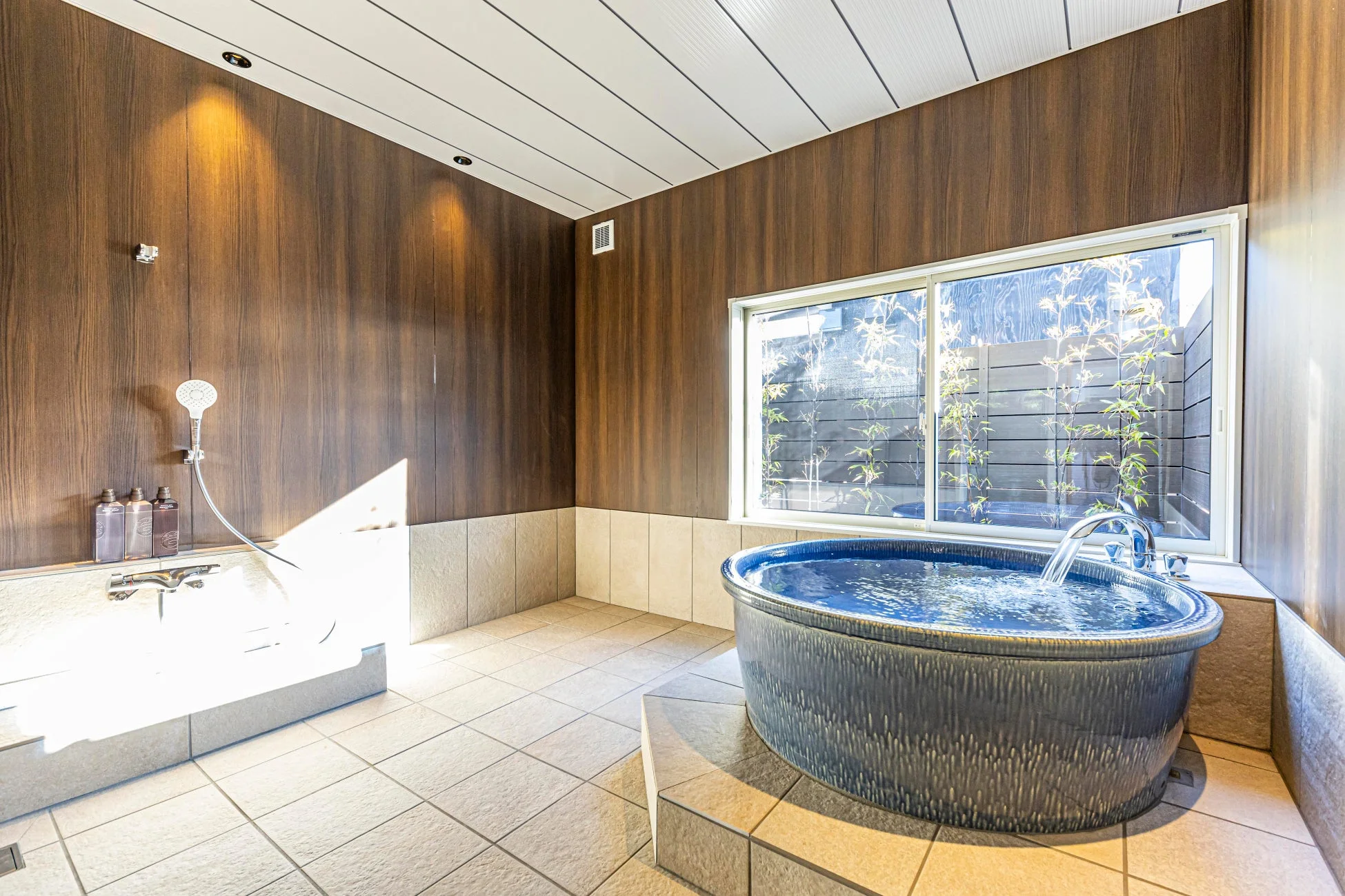 木目調の壁と明るいタイルで構成されたモダンな浴室に青い浴槽と白い浴槽が設置されている