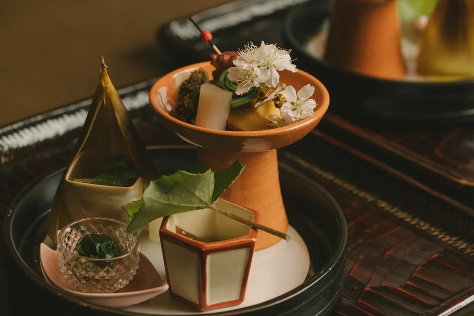 季節の食材が美しく盛り付けられた伝統的な和食の膳。桜のような花が添えられ、繊細で雅やかな懐石料理の一部を捉えた写真です。