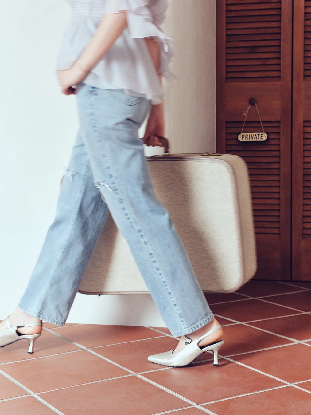 水色のダメージジーンズとフリルブラウスを着用した人物が、ヴィンテージ風のスーツケースを持ってテラコッタ調の床を歩いています。