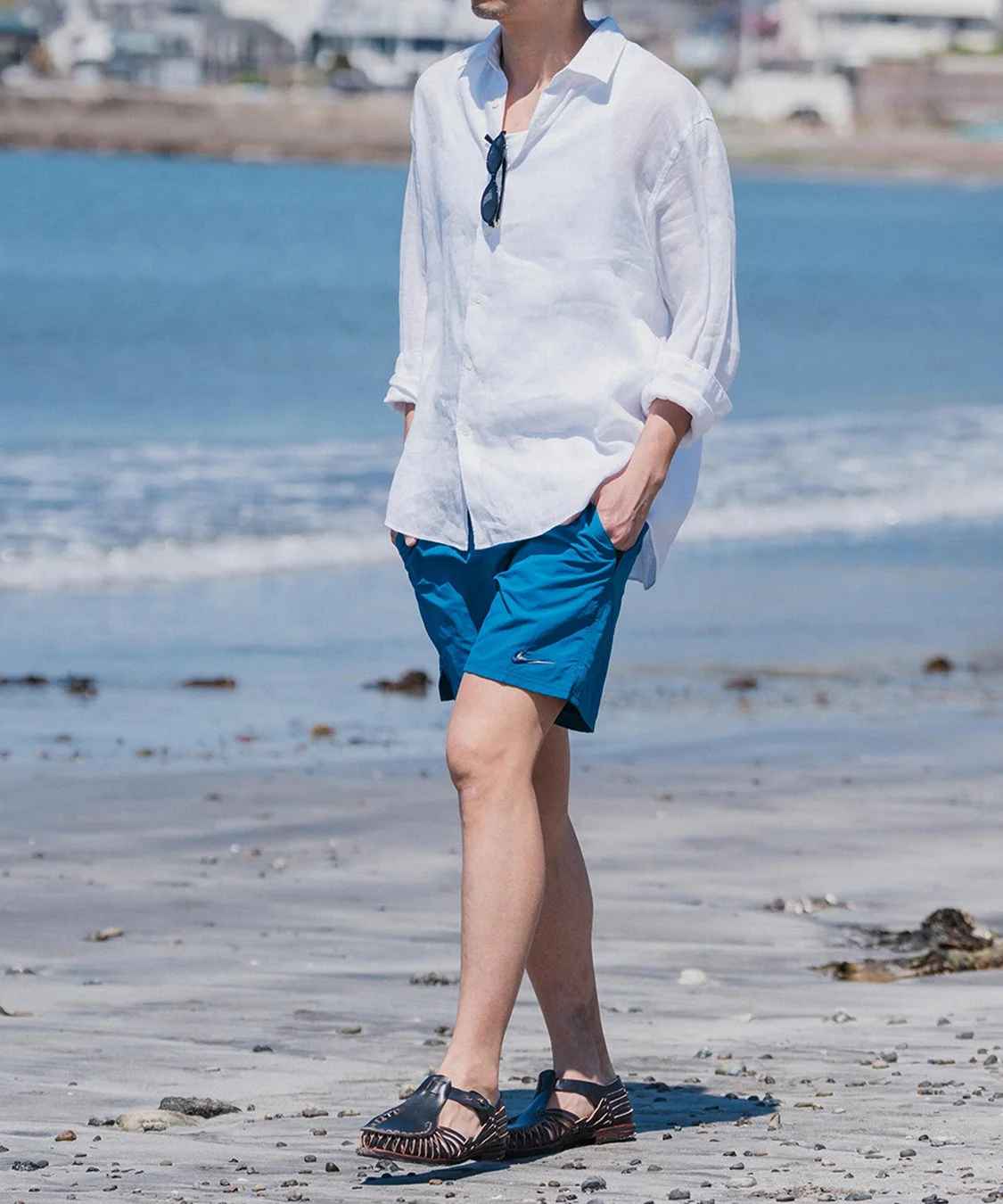 白いリネンシャツと青いハーフパンツを着用した男性がビーチに立っている様子。