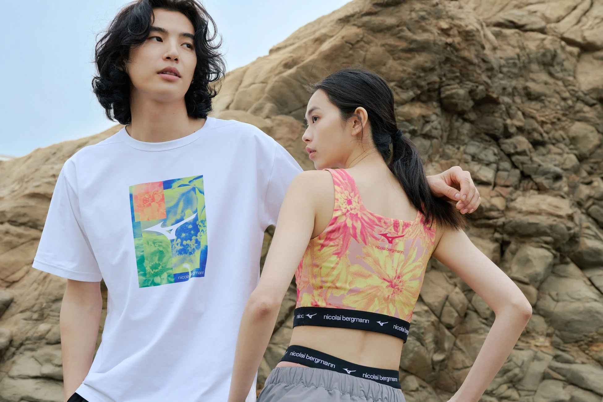 岩場を背景に、スポーツウェアを着た若い男女が並んで立つ姿を捉えた一枚。男性は白いTシャツ、女性は花柄のスポーツブラとショートパンツを着用。ニコライバーグマンとミズノのブランドロゴが見える。