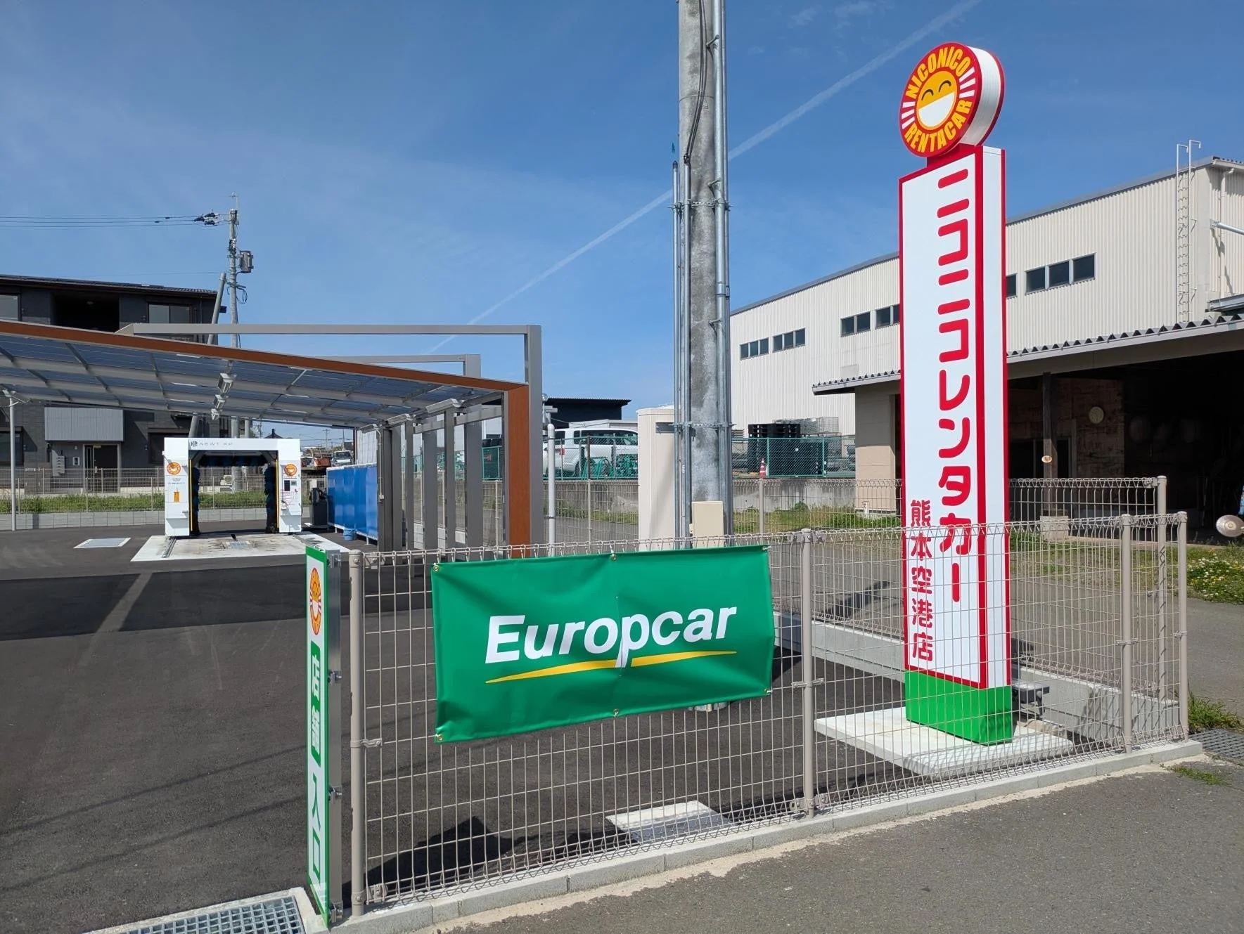 レンタカー会社の看板と洗車機が写っています。ニコニコレンタカーとEuropcarのサインが目立ち、熊本空港店の施設であることがわかります。