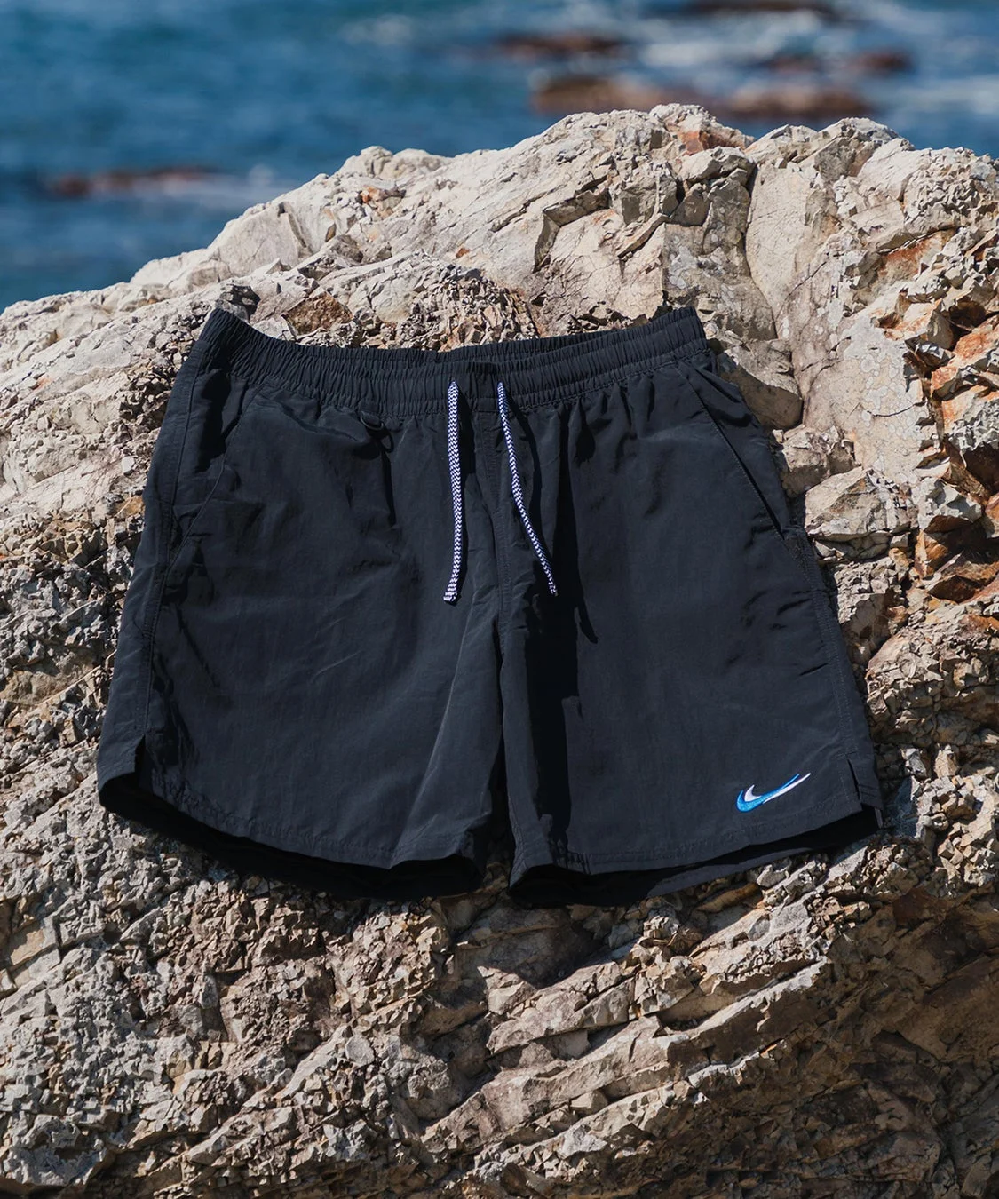 黒いショートパンツが岩場に置かれている写真です。パンツの裾には青い曲線状のロゴが見え、背景には青い海が広がり、夏のアウトドアやビーチでの着用を想起させます。