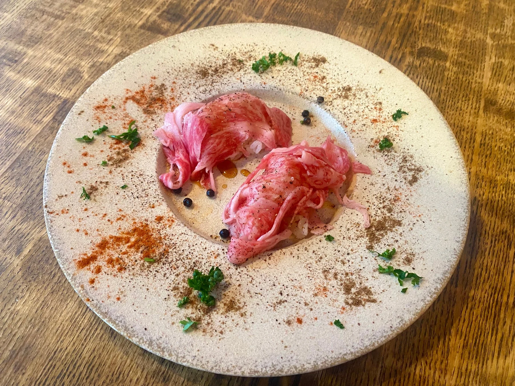 白い皿に盛り付けられた薄切りの生肉料理。肉は鮮やかな赤色で美しいサシが入っており、赤と黒のスパイス、そして緑のハーブが添えられ、見た目も華やかで食欲をそそる一品です。