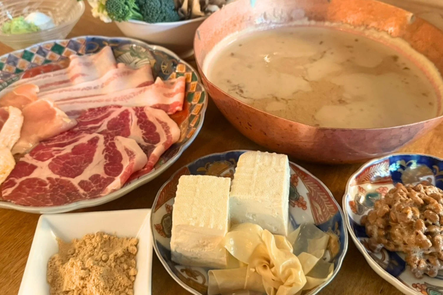 銅製の鍋を中心とした食卓で、薄切り肉、豆腐、湯葉、納豆、きな粉など様々な具材が並べられている