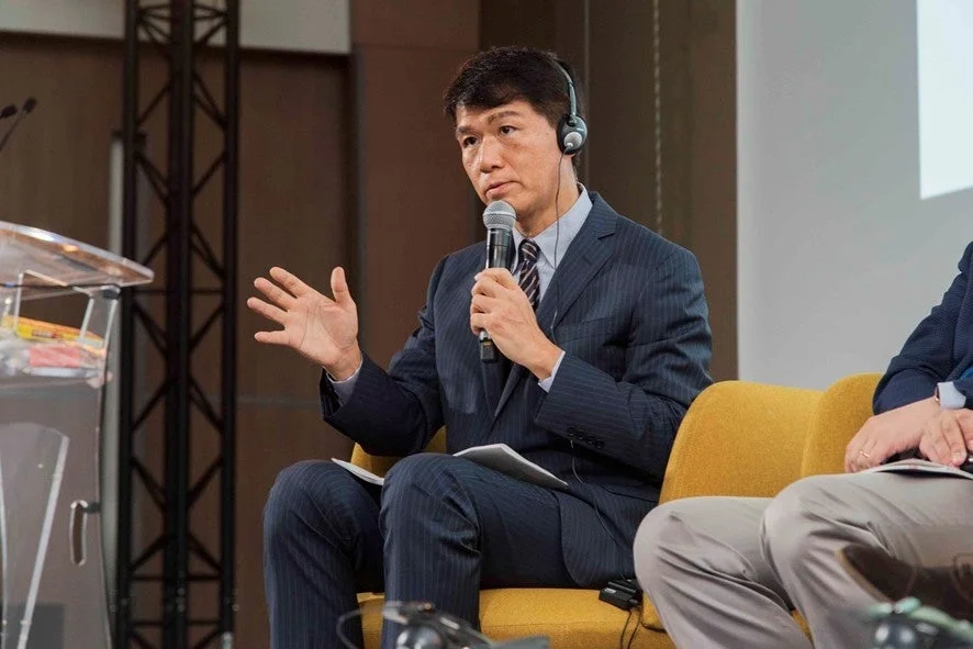 スーツを着たアジア人男性がヘッドホンを装着し、マイクを持って講演またはパネルディスカッションに参加している様子。