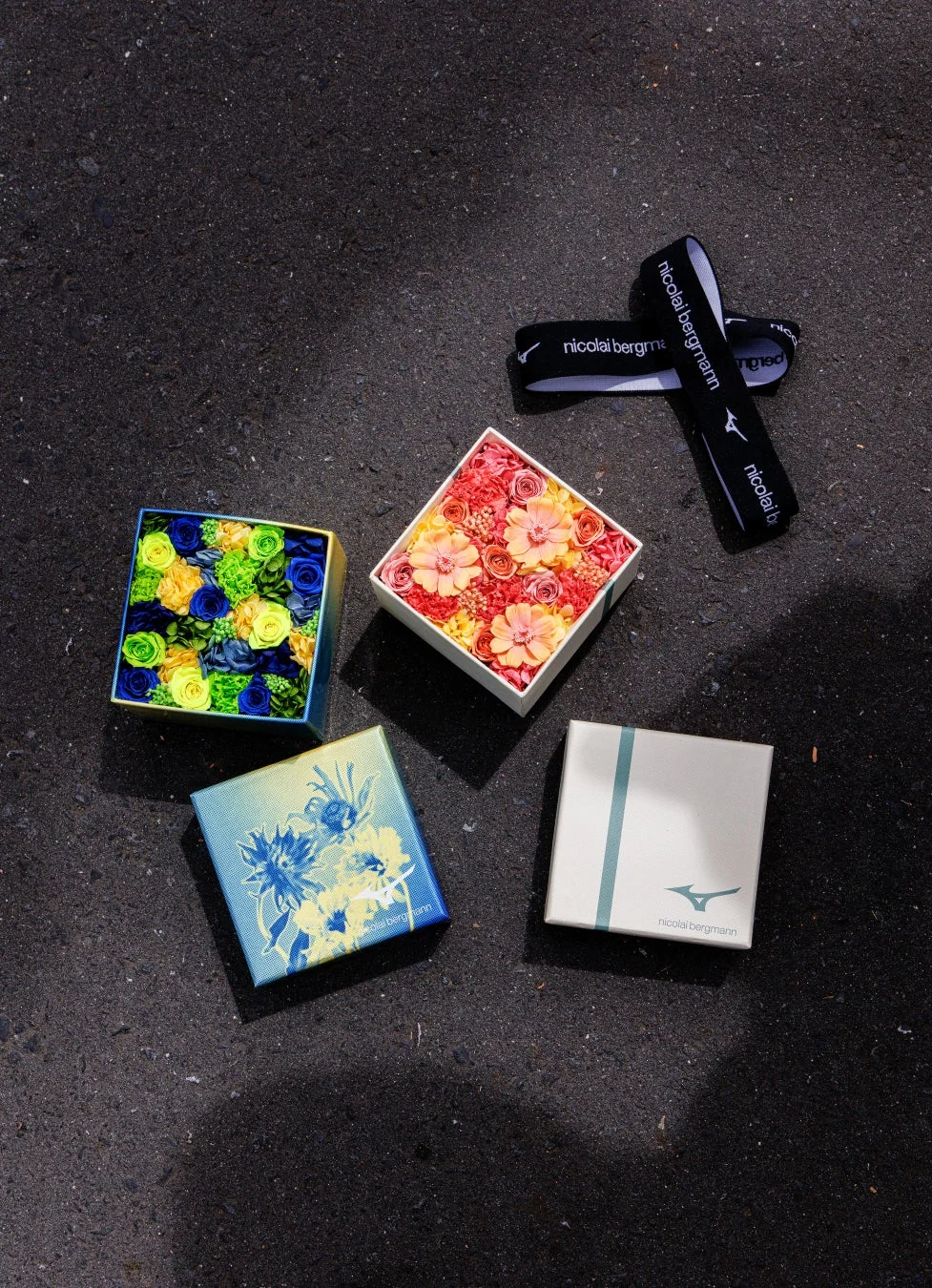 ニコライバーグマンとミズノのコラボレーションアイテムが、暗いアスファルトのような背景に並べられています。色鮮やかなプリザーブドフラワーのボックス、ブランドロゴ入りのギフトボックス、そしてロゴ入りバンドが見られます。