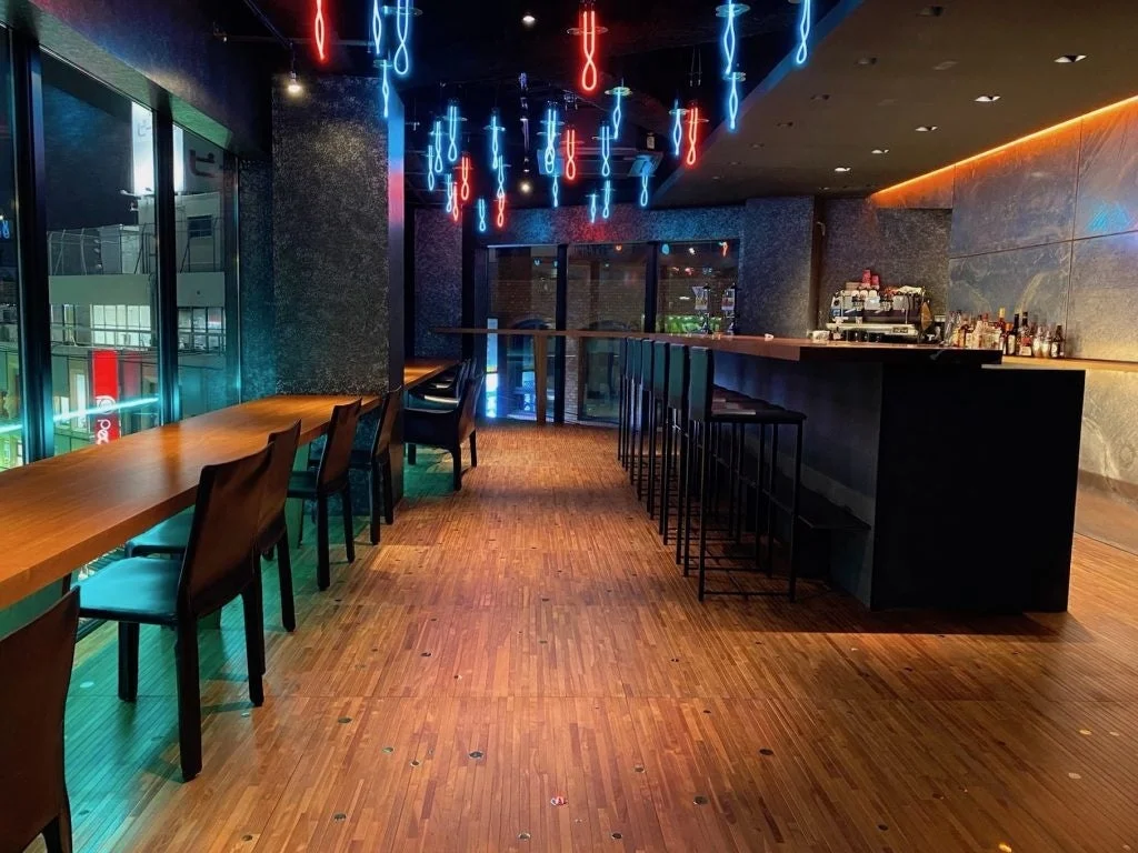 赤と青のネオンライトが印象的なモダンでスタイリッシュなバーの店内。窓からは夜景が広がり、木製のカウンター席とバーカウンターが並びます。落ち着いた照明が大人の雰囲気を演出しています。