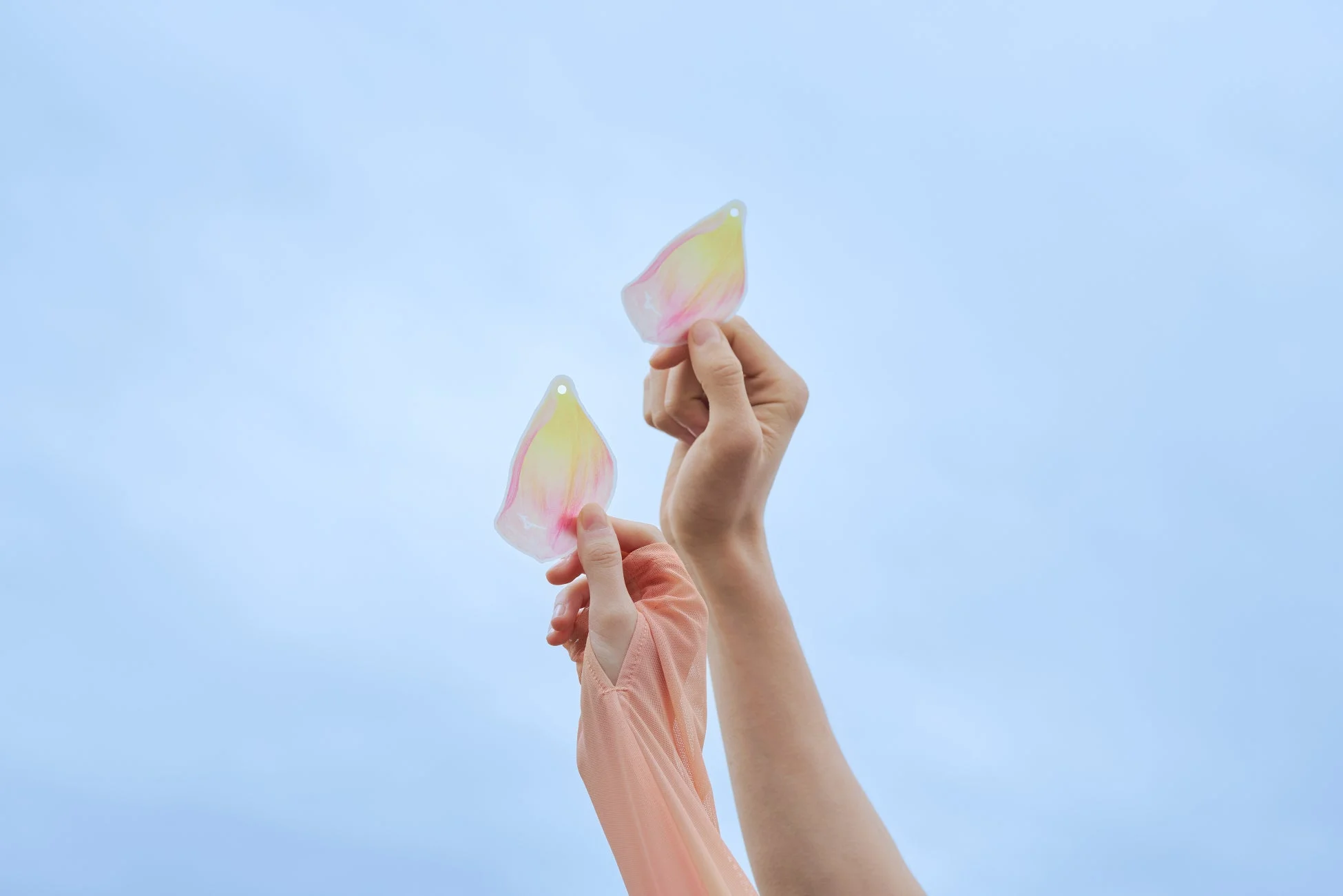 青空を背景に、2つの手が淡いピンクと黄色のグラデーションが施された透明な花びら型の飾りを掲げています。軽やかで繊細な美しさを感じる一枚です。