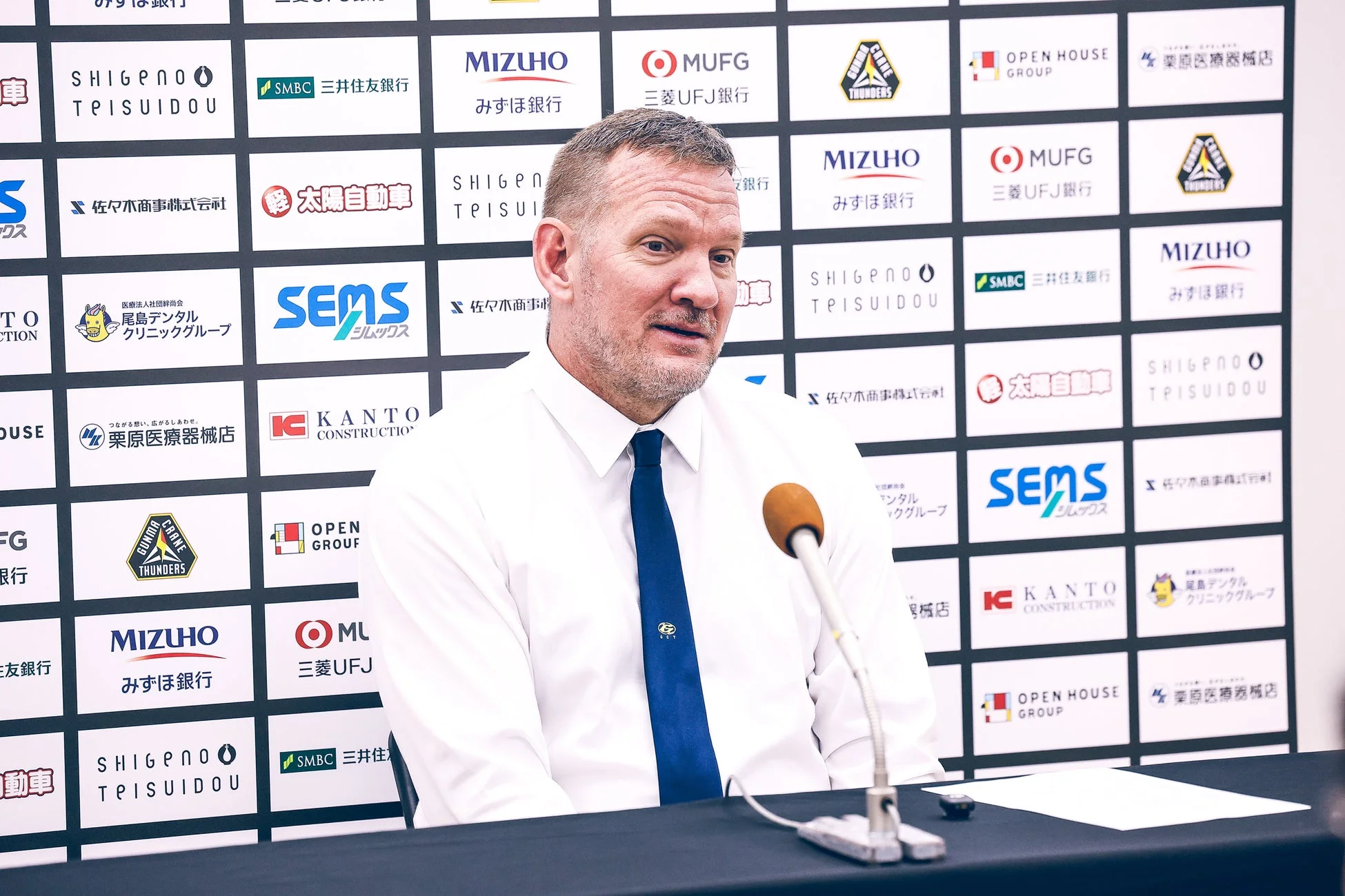 白いシャツと青いネクタイを着た男性が、スポンサーロゴが並ぶ背景の前でマイクに向かって話している様子