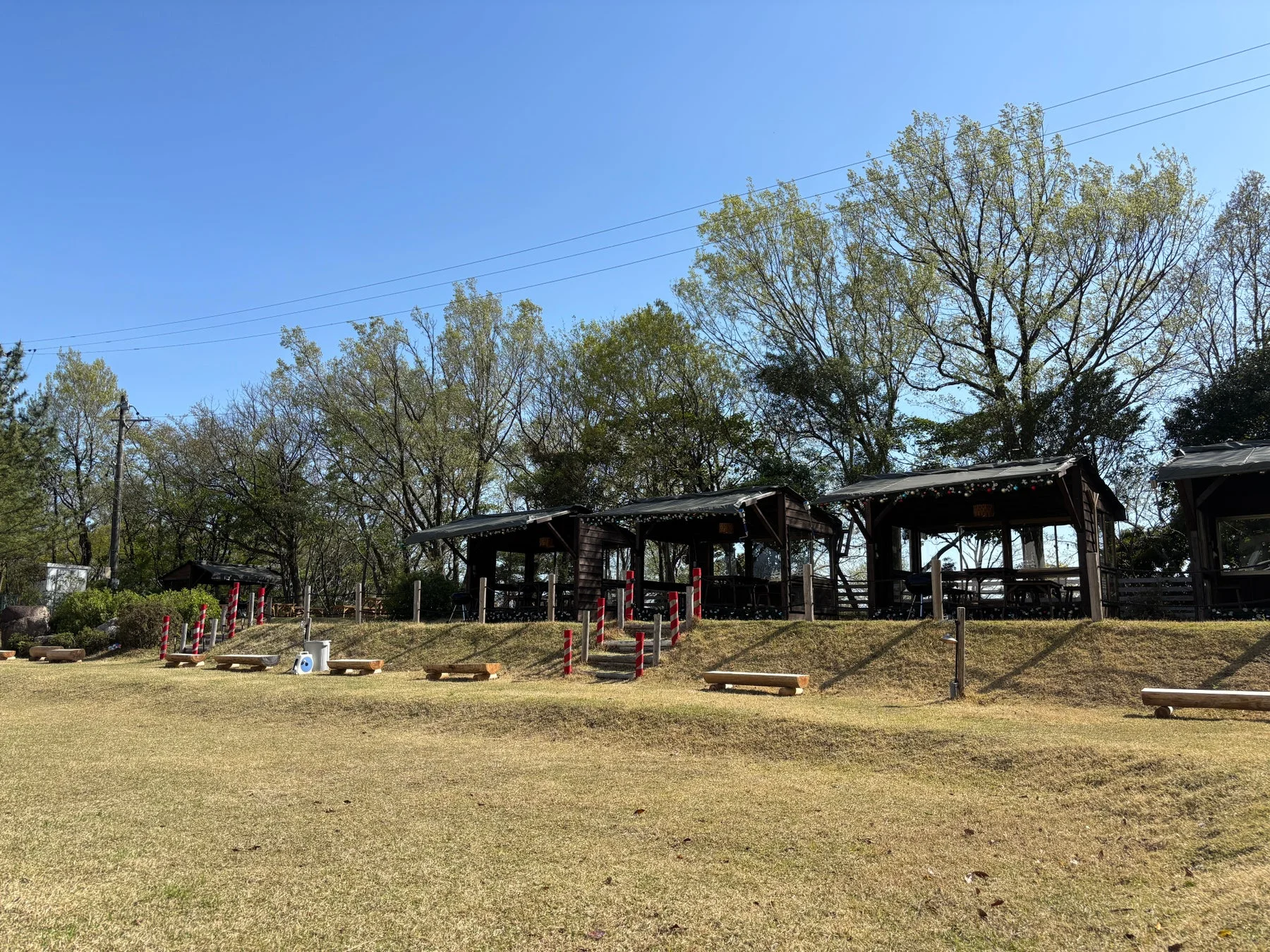 晴れた日の屋外のレジャー施設を捉えた画像。芝生の広場と木製の屋根付き休憩所が並ぶ