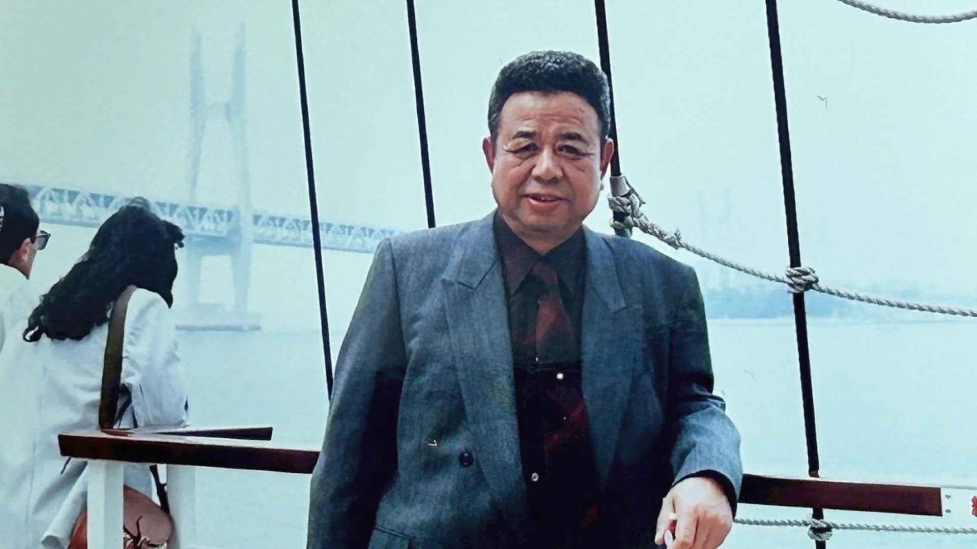 船上と思われる場所で、スーツを着た中高年男性がカメラに向かって立っている。