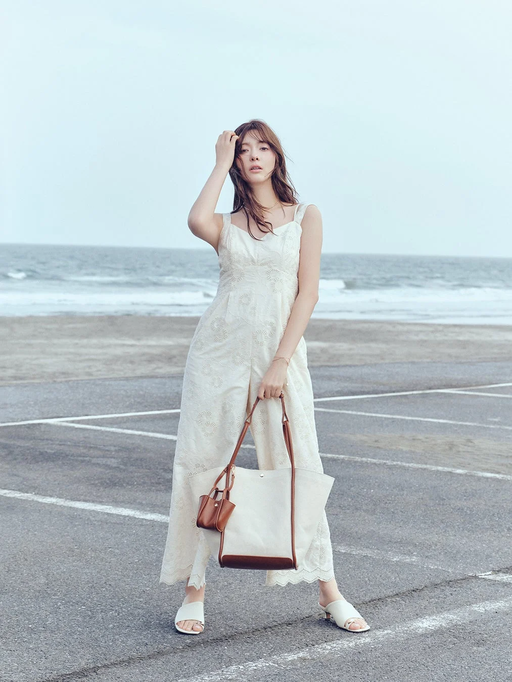 海辺の駐車場で、白いレースのオールインワンを着た女性がポーズをとっています。