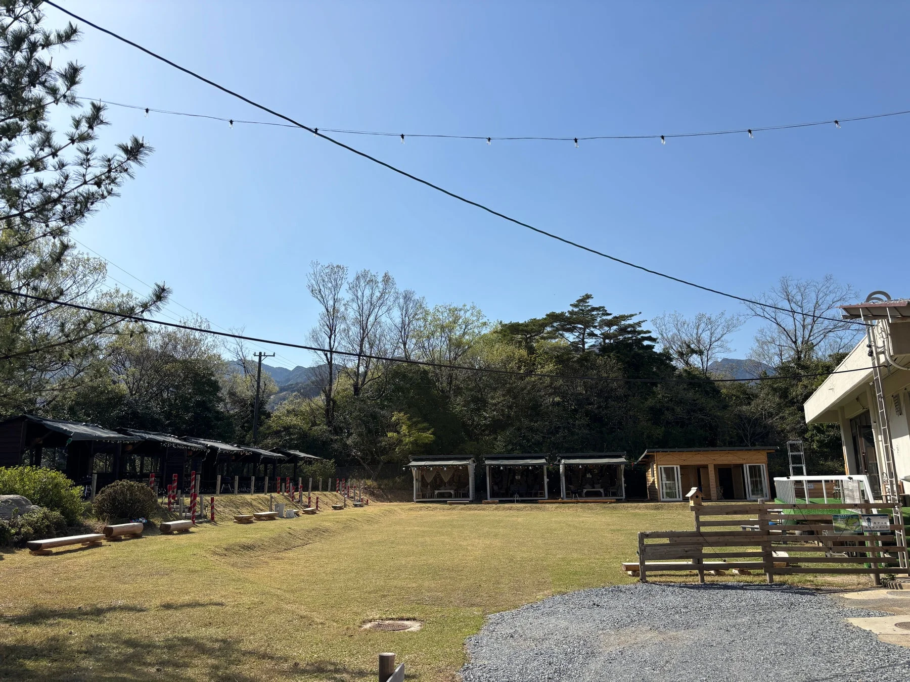 晴れた日に広がる芝生の広場と、周りを囲む木々や山々、そして小さな休憩所が見えるアウトドア風景