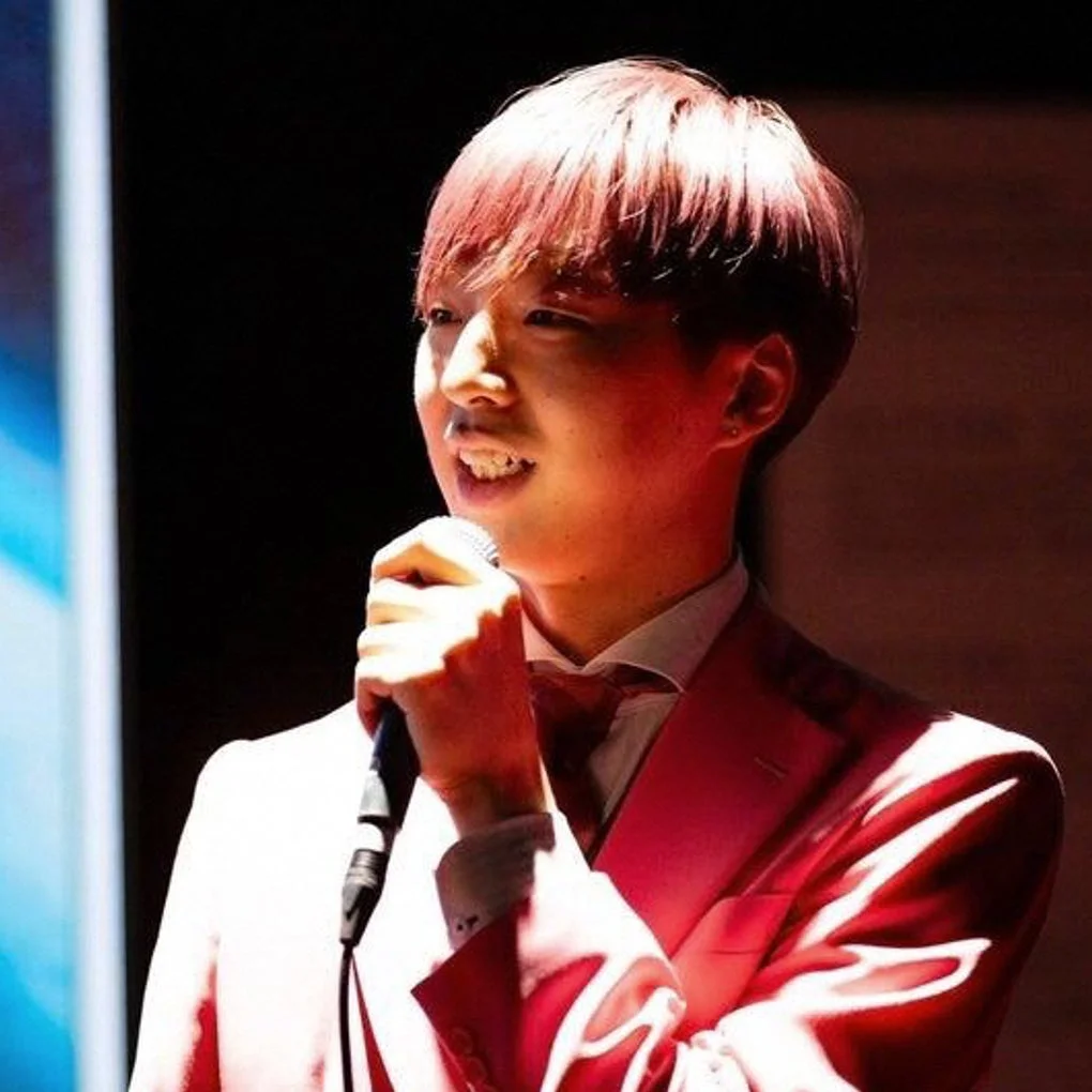 ピンクのスーツと蝶ネクタイを着用した若い男性がステージ上でマイクを持って歌唱またはスピーチをしている