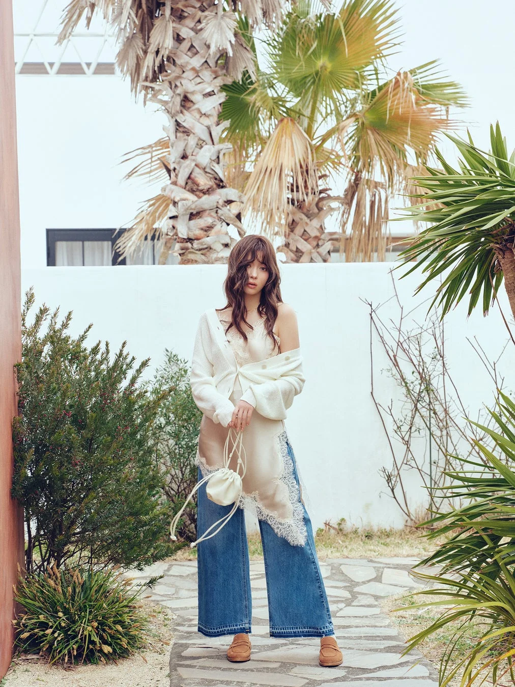 白いカーディガンとレースのスリップドレス、ワイドデニムを着用した女性が、パームツリーのある屋外でポーズをとっています。