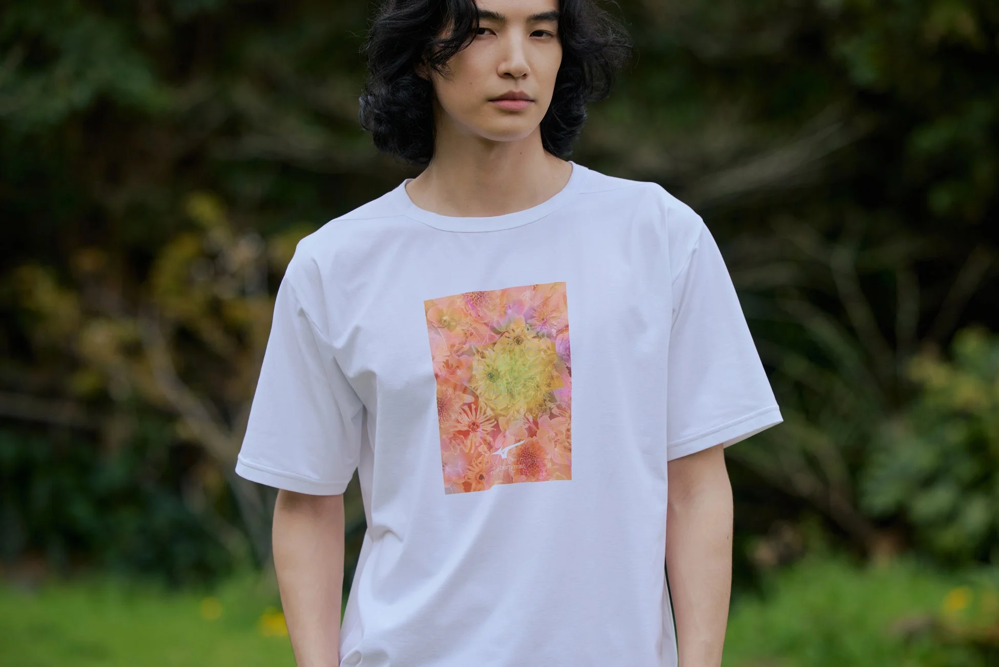 長い黒髪の男性が白いTシャツを着て立っています。Tシャツにはピンクや黄色の花柄がプリントされており、背景は緑豊かな自然です。若々しくカジュアルな印象のポートレート写真です。