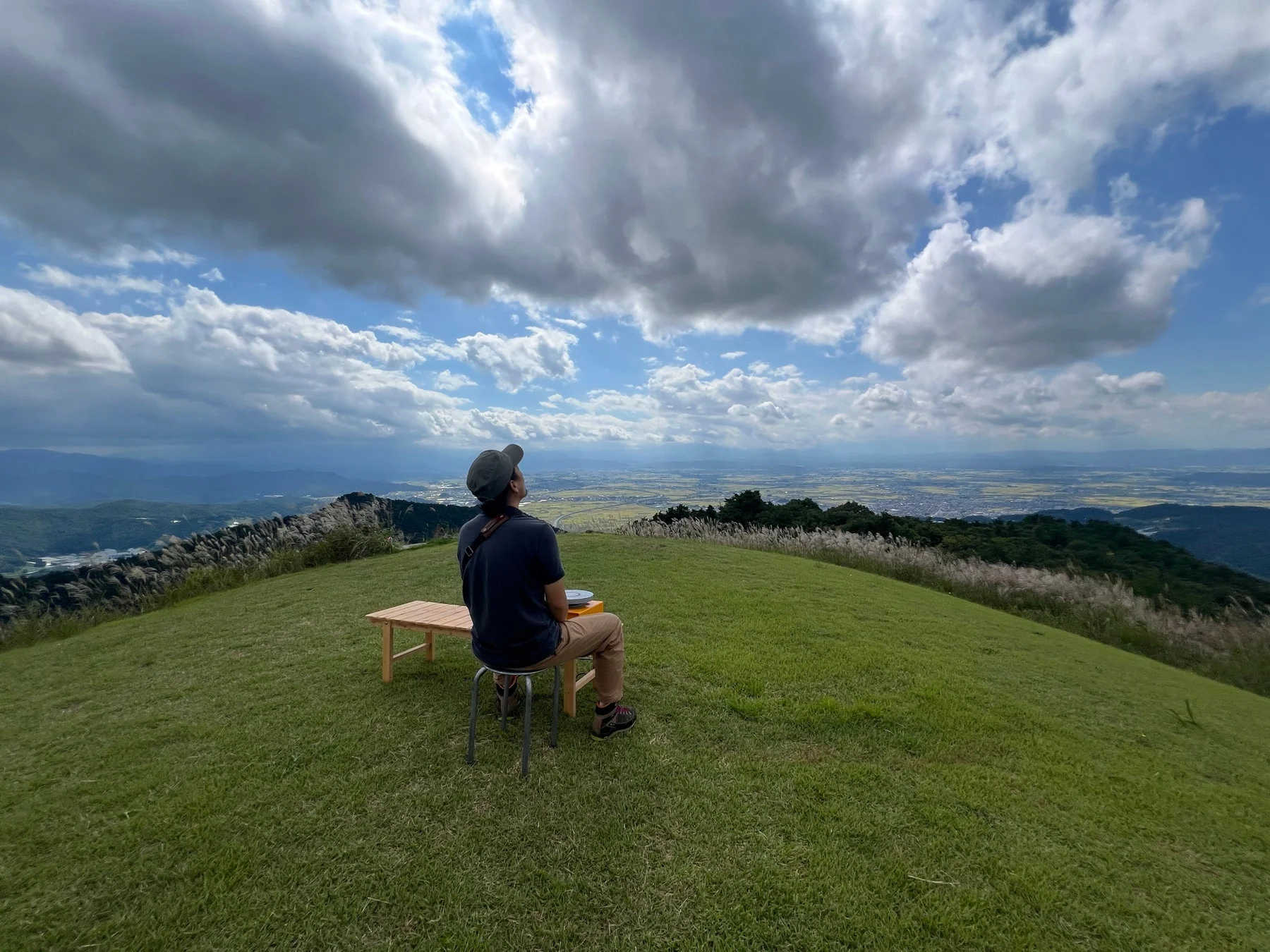 緑豊かな丘の頂上に座る男性が、広がる雲と遠くの景色を眺めている