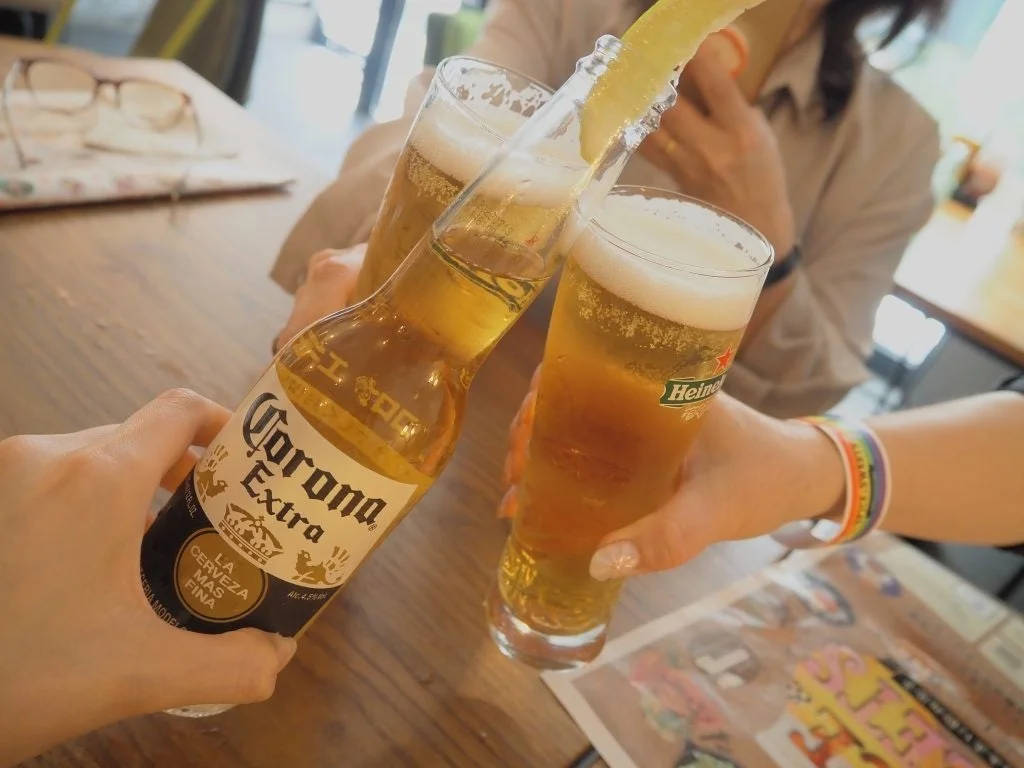 友人たちが集まってビールで乾杯している瞬間を捉えた写真です。コロナビールにはライムが添えられ、ハイネケンのグラスもあります。楽しい雰囲気の飲み会の様子が伝わってきます。