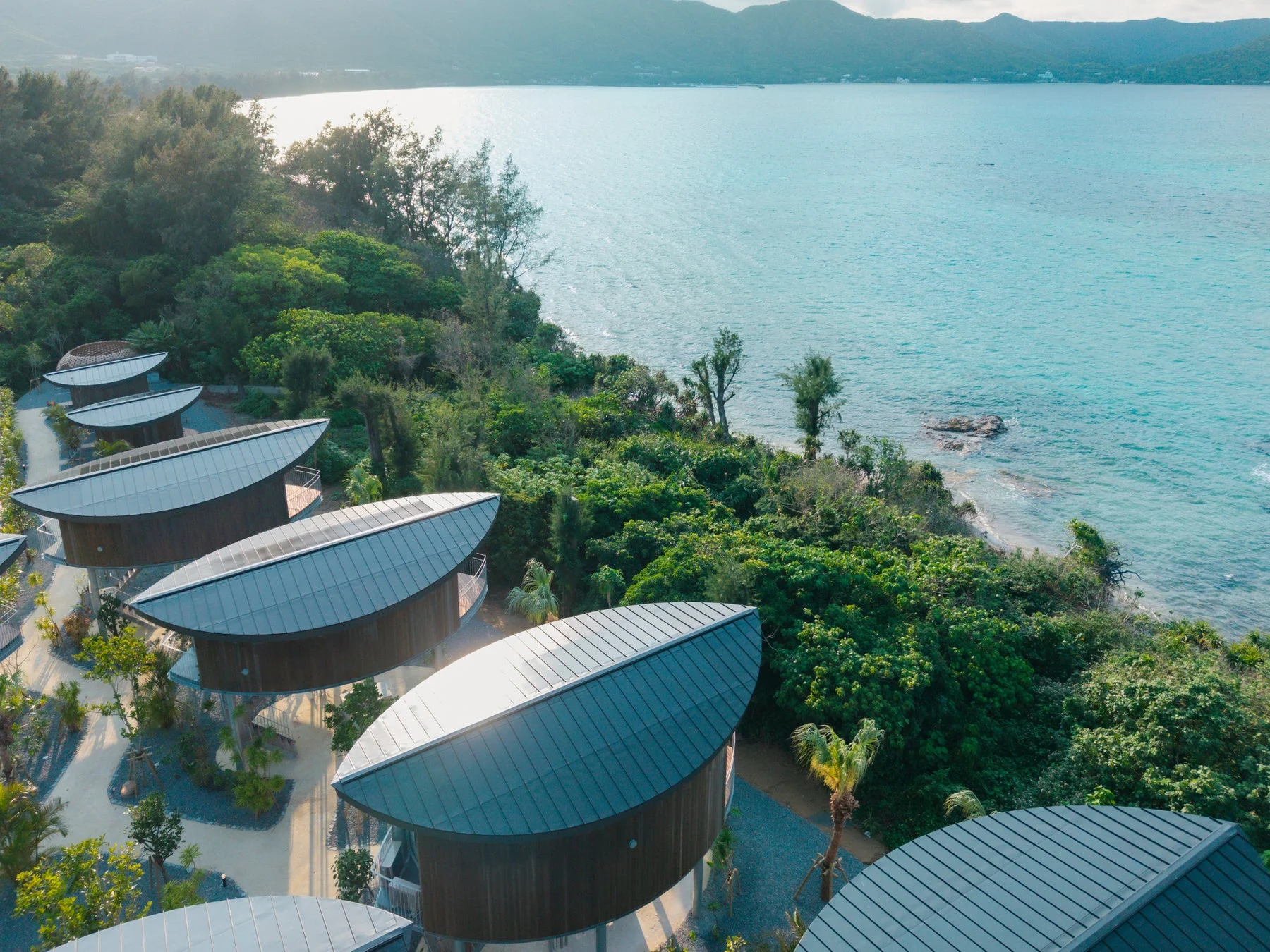 曲線的な屋根を持つモダンな建物群が、豊かな緑に囲まれ青い海に面して建ち並ぶリゾート地の空撮