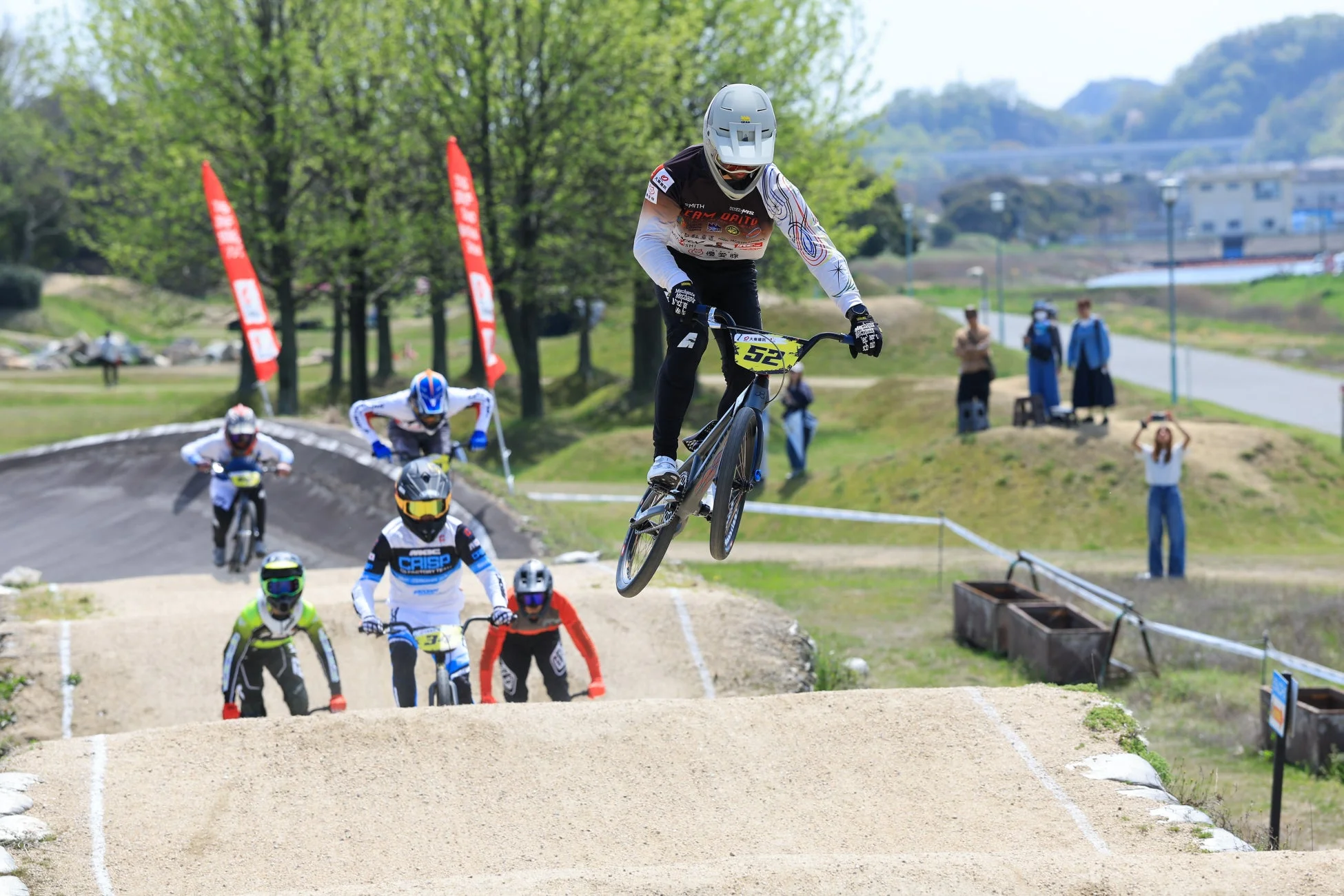 BMXレースで選手たちがダートトラックを疾走し、一人の選手が大きくジャンプしている瞬間を捉えた写真