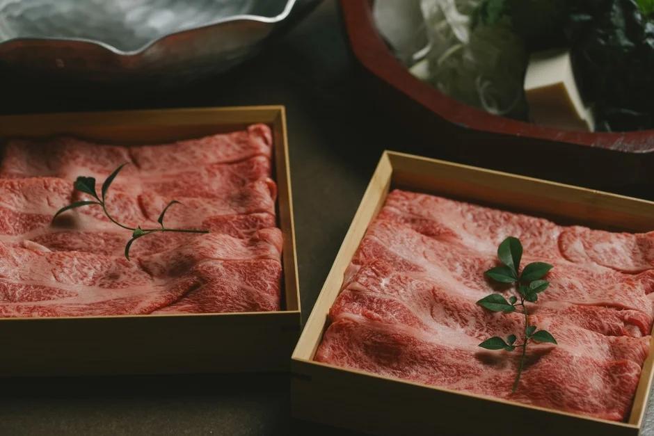 木箱に美しく盛り付けられた霜降り和牛の薄切り肉が二箱並べられています。緑の葉が添えられ、背景には鍋料理の具材が見え、高級感あふれる食卓の準備が伺えます。