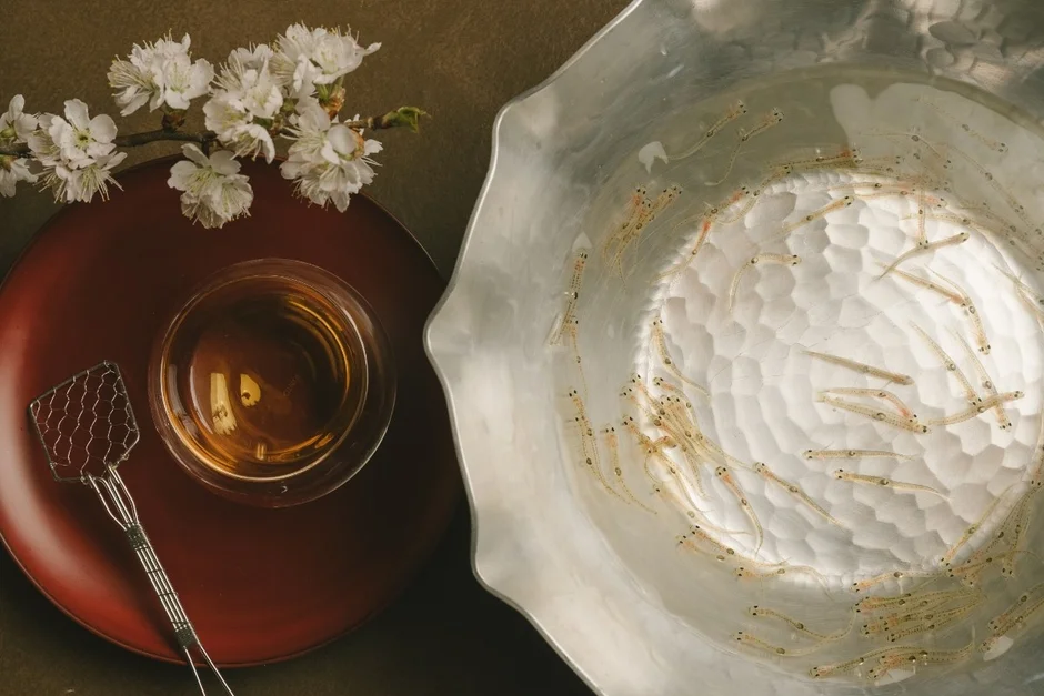 たくさんの白い小魚(しらす)が入った銀色の器と、赤い漆器の皿に置かれた飲み物(お茶か酒)、そして小さな網杓子が写っています。白い花が添えられ、新鮮な食材と日本の食卓の風情を感じさせる一枚です。