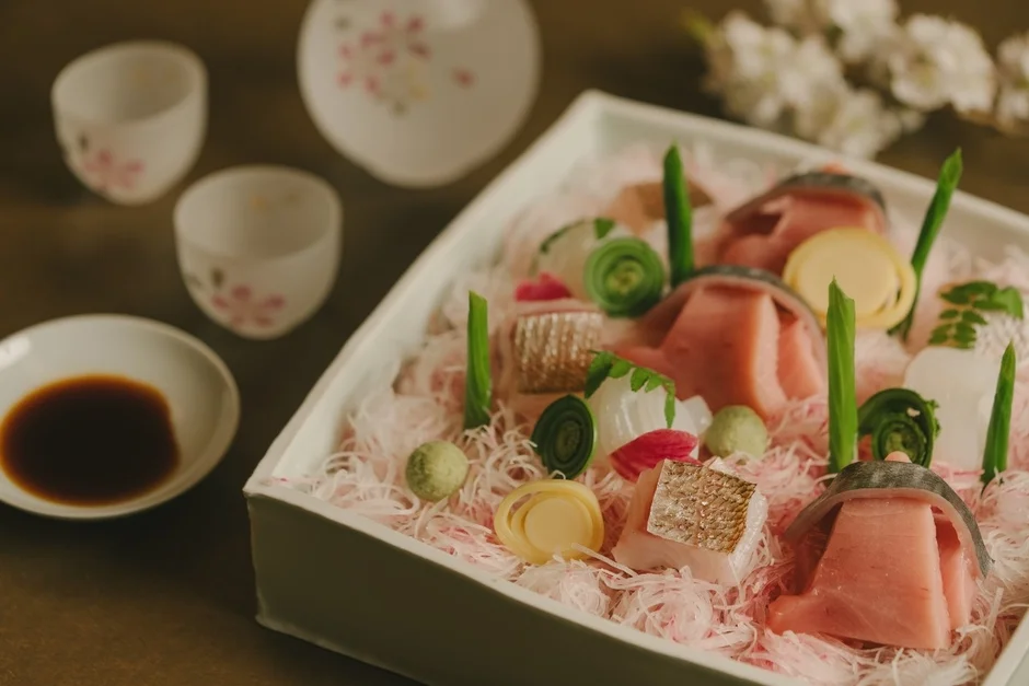 豪華な刺身の盛り合わせが白い箱に美しく盛り付けられています。マグロやアワビなど様々な種類の新鮮な魚介が並び、背景には桜柄の日本酒の器や花が見え、上品で特別な食事の風景を示唆しています。