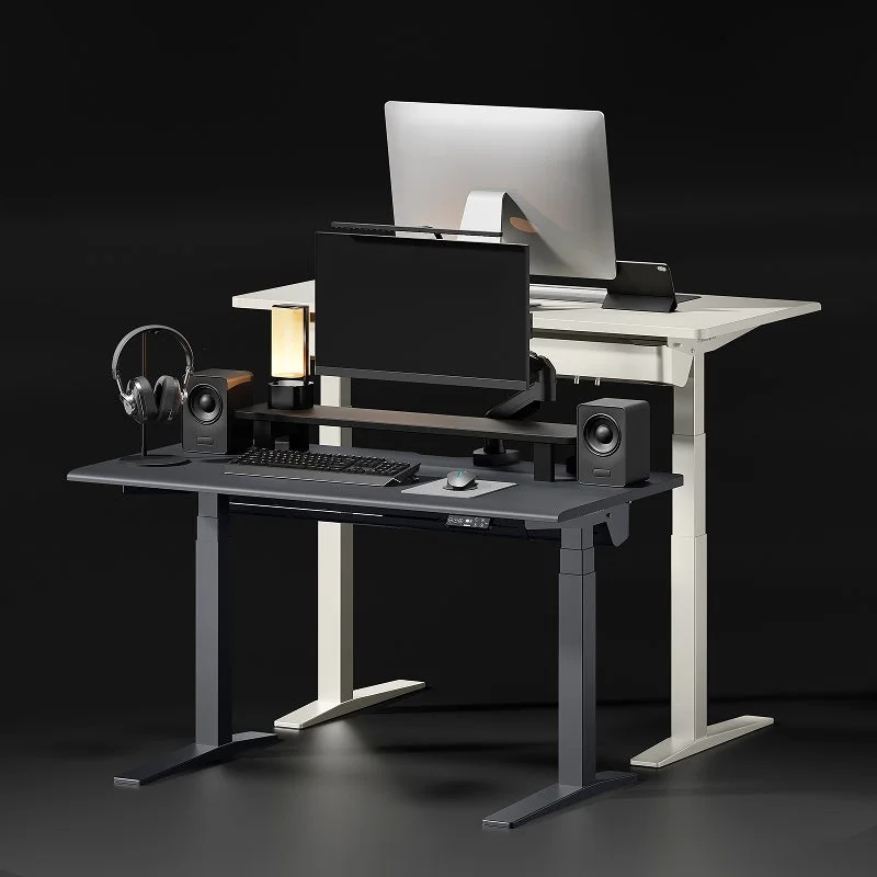 2台の昇降式デスクが並べられており、手前のダークグレーのデスクにはモニター、キーボード、マウス、スピーカー、ヘッドホンなどが配置されている。奥の白いデスクも同様にモニターが置かれ、現代的なワークスペースを構築している様子。
