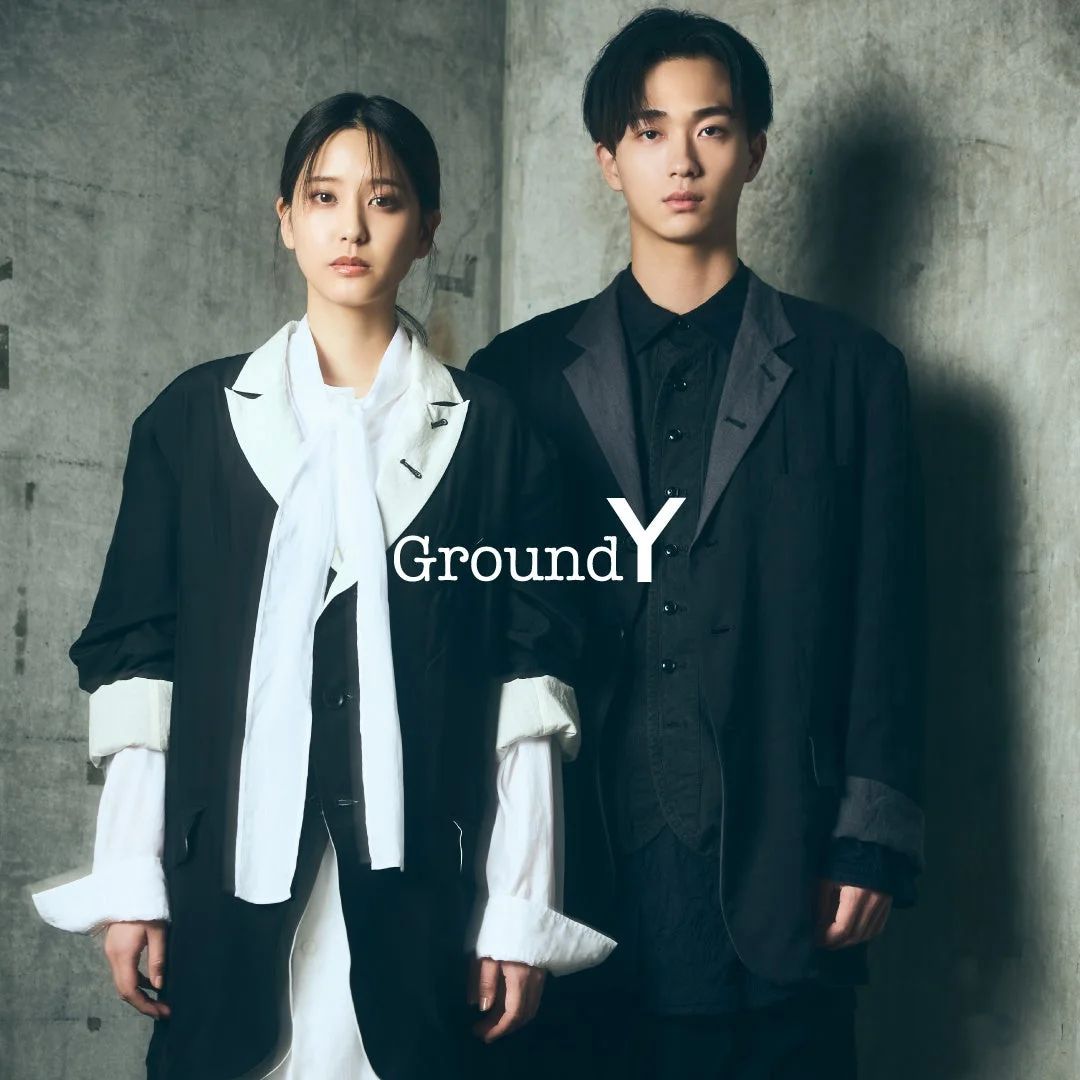 若い男女モデルが「Ground Y」のモダンなモノクロファッションを着用し、コンクリート壁を背景にポーズをとっています。
