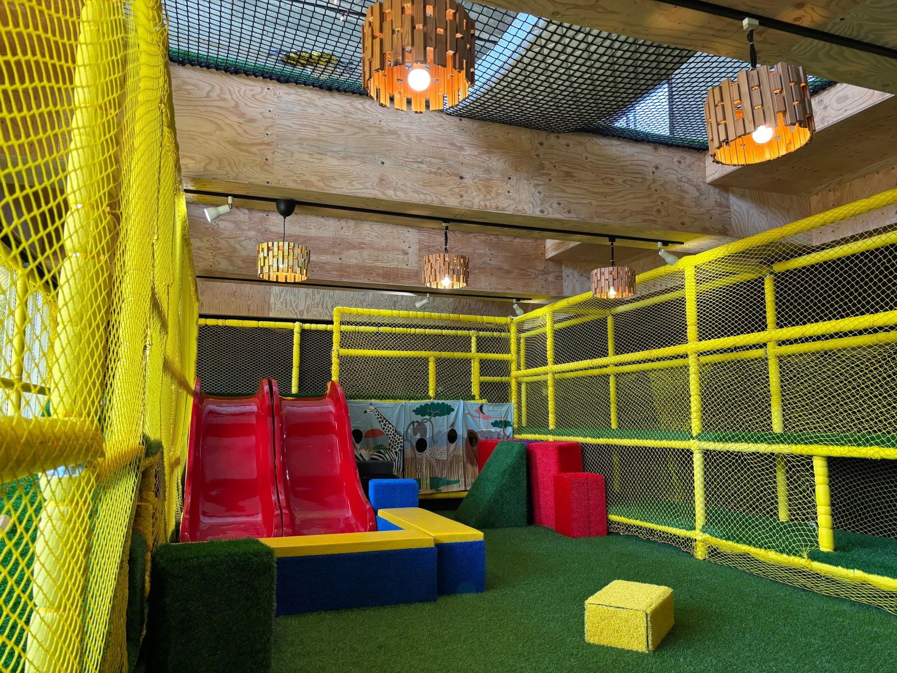 黄色いネットで囲まれた室内遊び場。赤い滑り台や様々な形状のソフトブロックが配置され、床は人工芝で覆われている