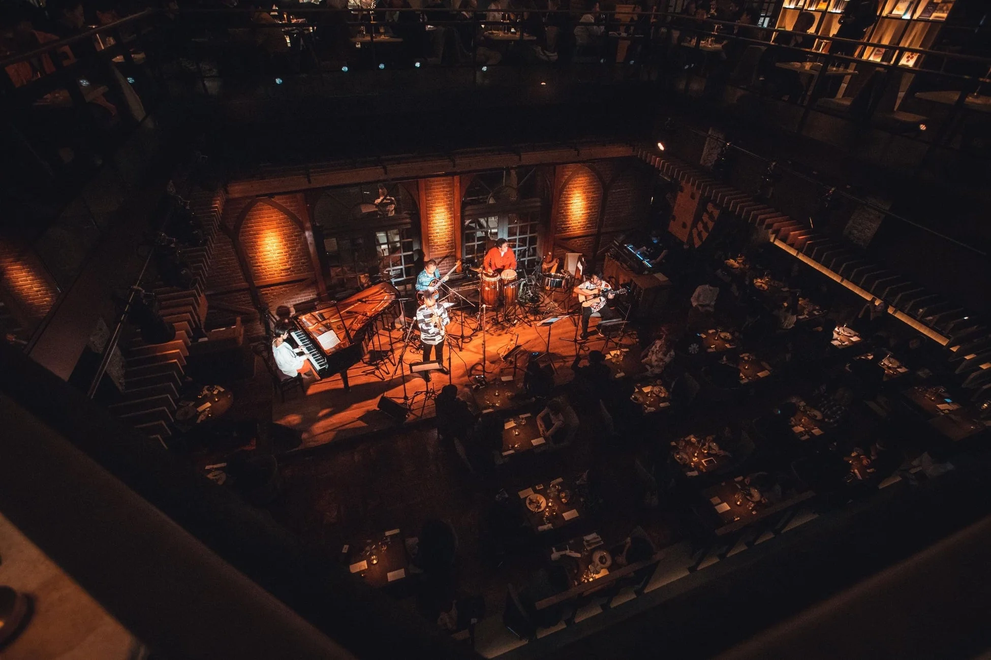 薄暗い空間でバンドがライブ演奏を行っており、多くの観客が食事やドリンクを楽しみながら音楽に耳を傾けている様子