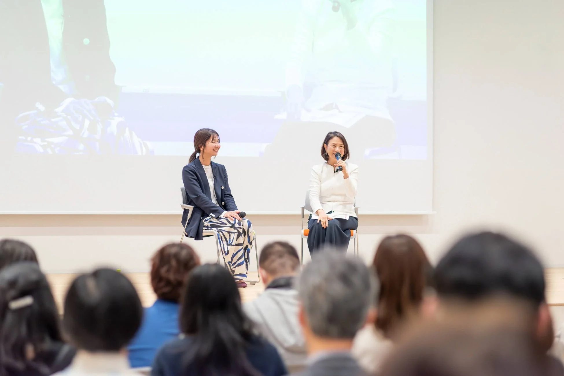 ステージ上で二人の女性が座っており、一人がマイクを持って話しています。もう一人の女性は笑顔で聞き入っており、手前には聴衆の後ろ姿がぼんやりと写っています。