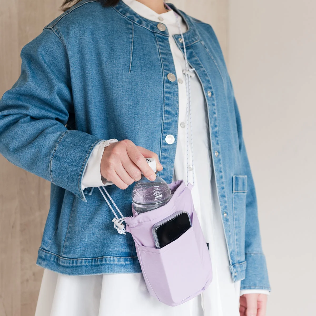 デニムジャケットと白のワンピースを着用した人物が、薄紫色のショルダーバッグを持ち、中に入っているペットボトルを手にしている様子を捉えた画像です