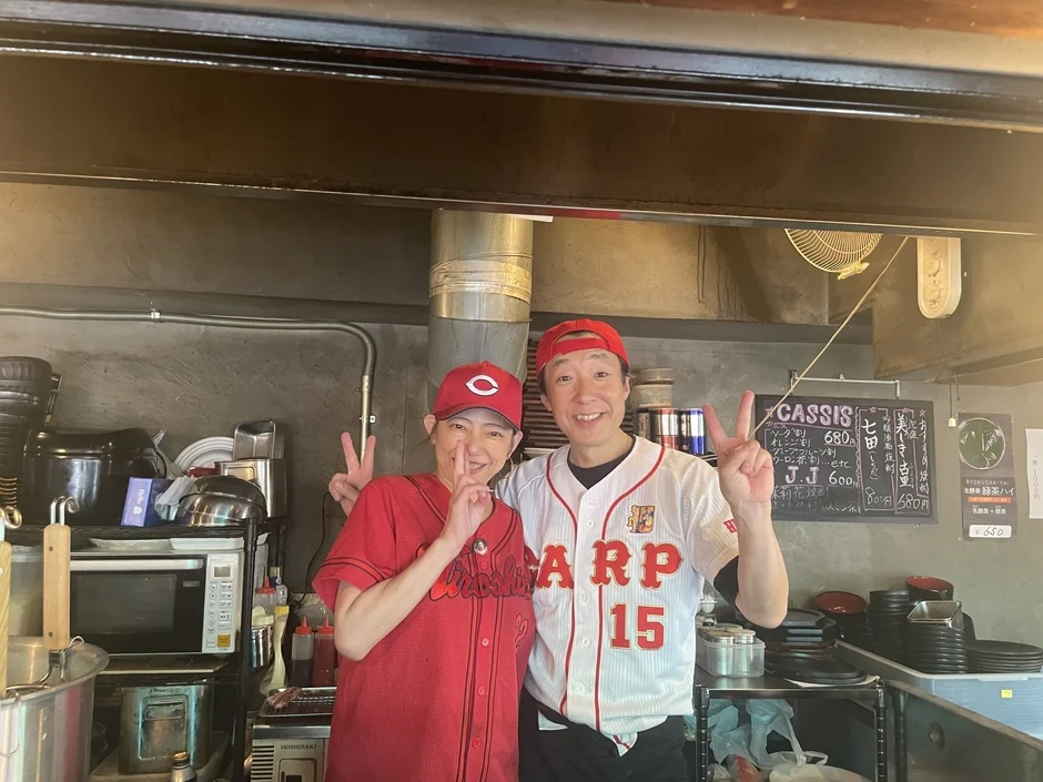 広島東洋カープのユニフォームとキャップを着用した男女が、飲食店のカウンターで笑顔でピースサインをしています。