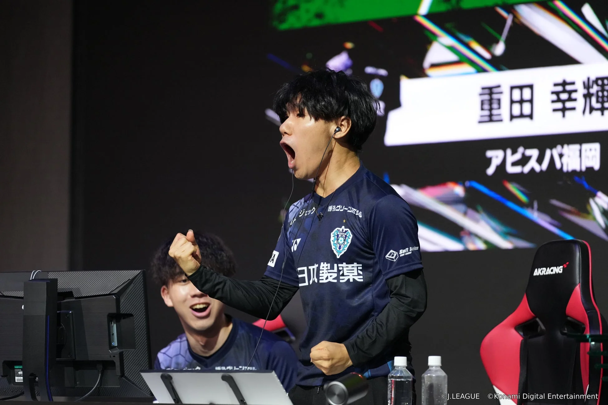 eスポーツの試合中に、青いユニフォームを着た男性選手が拳を握りしめ、口を大きく開けて感情を爆発させている瞬間