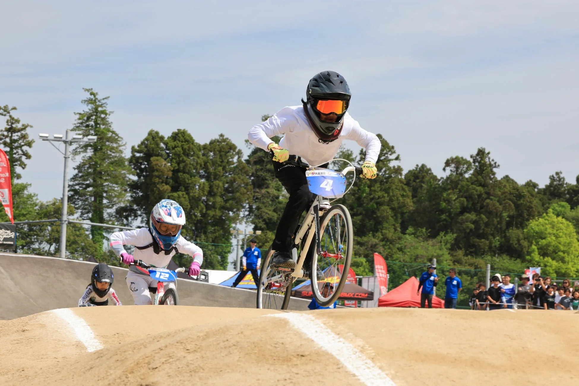 BMXレースでライダーたちがダートコースを疾走する躍動感あふれる瞬間。先頭のライダーはジャンプしており、ヘルメットとゴーグルを着用した別のライダーが続く。青空の下、競技の熱気が伝わる。