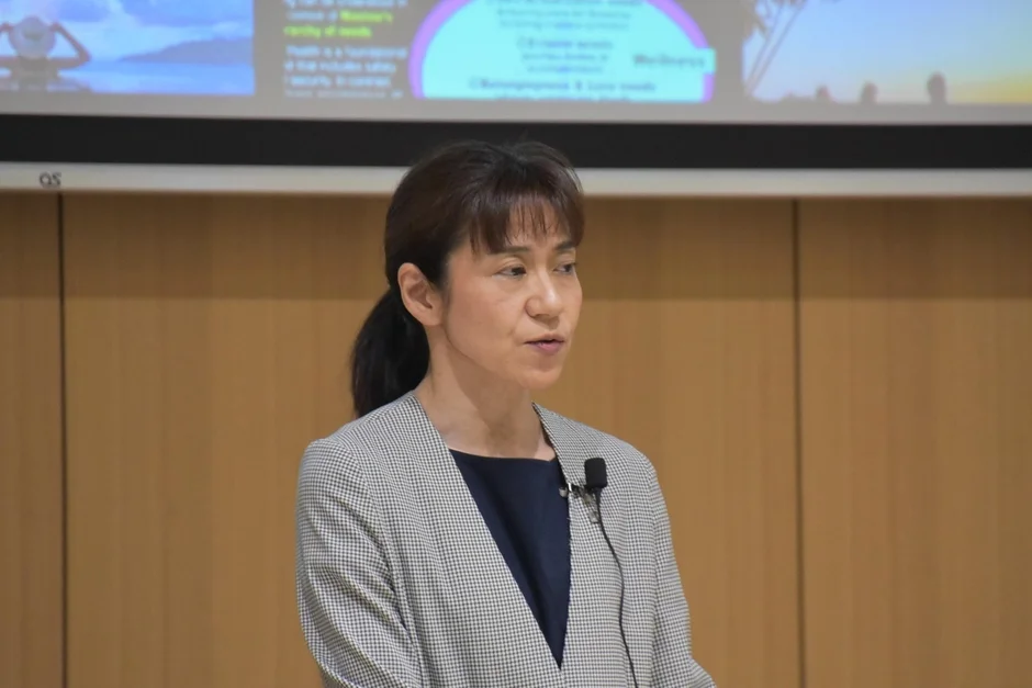チェック柄のジャケットを着てマイクを付けた女性が、講演またはプレゼンテーションを行っています。