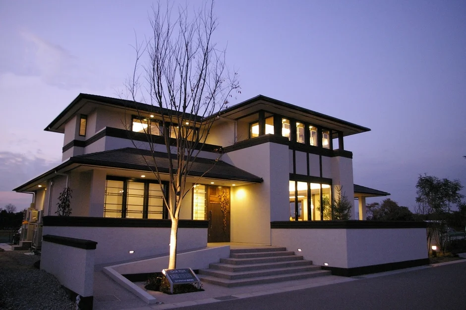 夕暮れ時、暖かく照明が灯されたモダンなデザインの一戸建て住宅の外観です。白とダークカラーのコントラストが特徴で、玄関前には裸の木が植えられ、窓から光が漏れています。住宅設備会社の看板も確認できます。