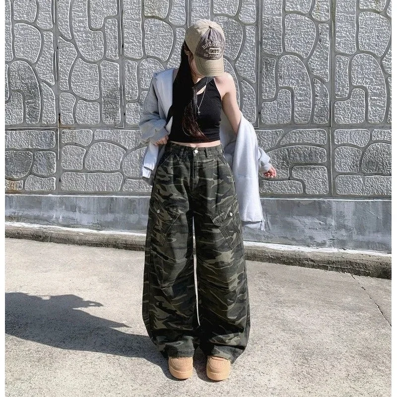 迷彩柄のカーゴパンツと黒のクロップトップを着用し、キャップをかぶった女性が壁の前でポーズをとっています。