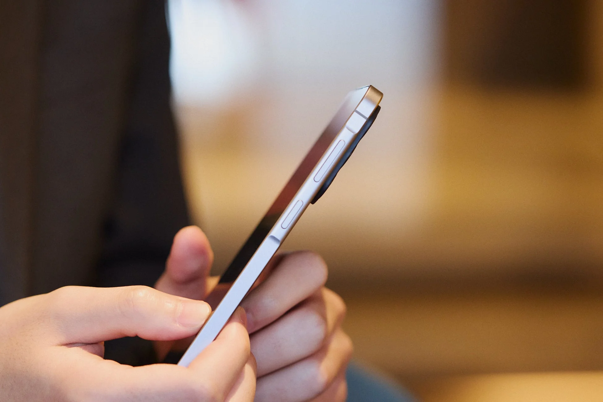 スマートフォンを操作している人物の手元を側面から捉えた画像です。デバイスの薄い側面と音量ボタンなどが鮮明に写っており、背景はぼかされています。