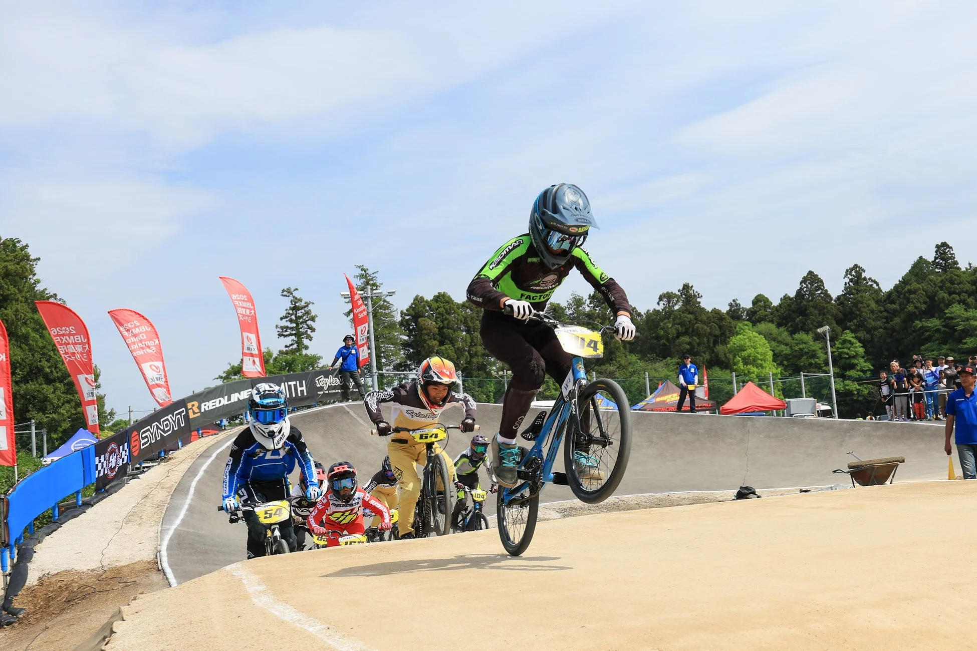 青空のもと、複数のBMXレーサーがダートコースで激しいレースを繰り広げている。先頭の選手が自転車で高くジャンプし、他の選手たちも続く。コース脇には観客やスタッフ、スポンサーの旗が見える。