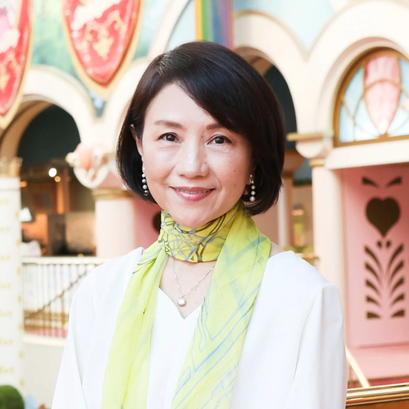 白いトップスと緑色のスカーフを身につけた笑顔の東洋人女性のポートレートです。パールイヤリングとネックレスを着用し、背景にはカラフルで装飾的な建物が広がっています。