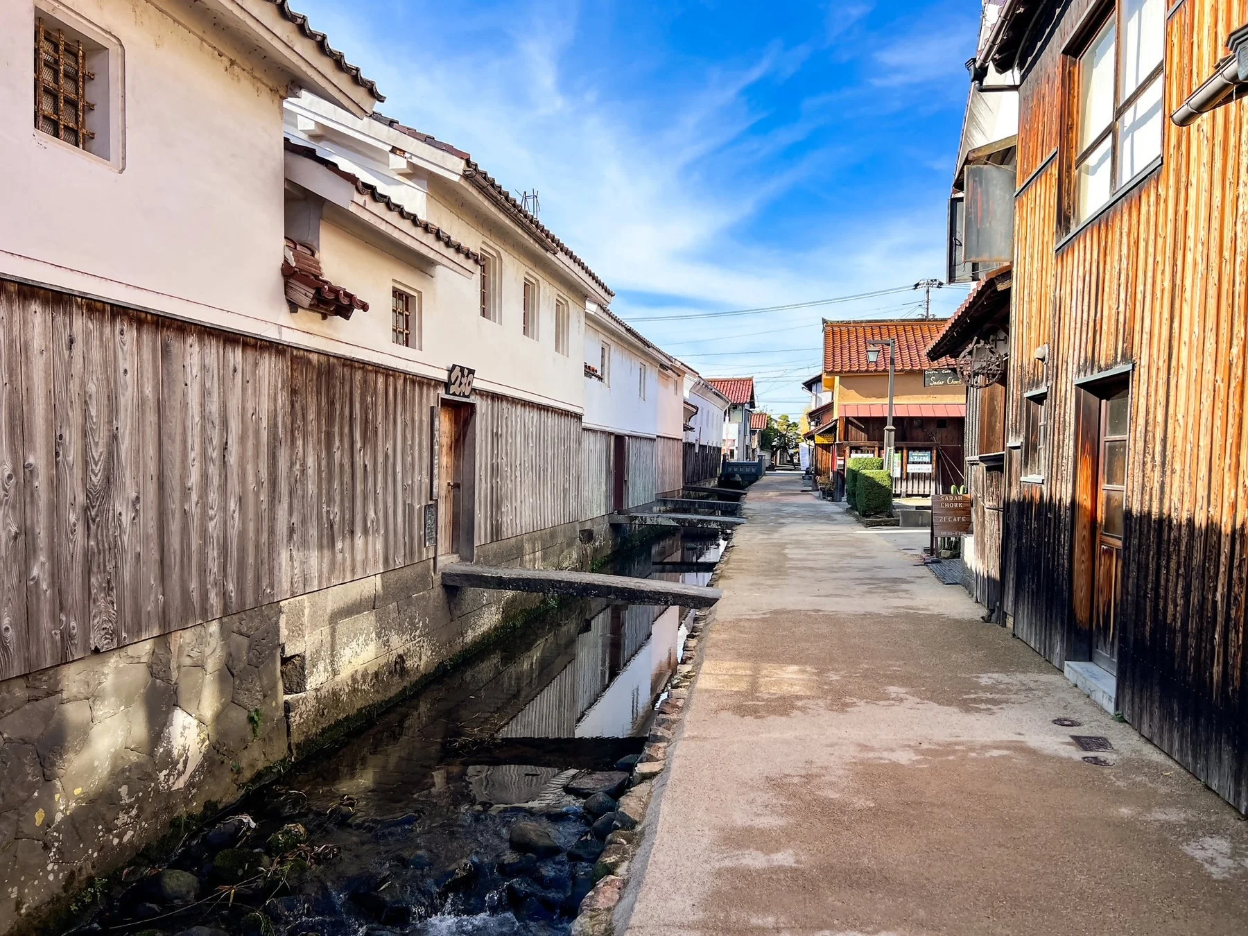 青い空の下、伝統的な日本家屋が立ち並ぶ古い街並みを捉えた画像です。家屋の傍らには水路が流れ、趣のある小橋が架けられています。