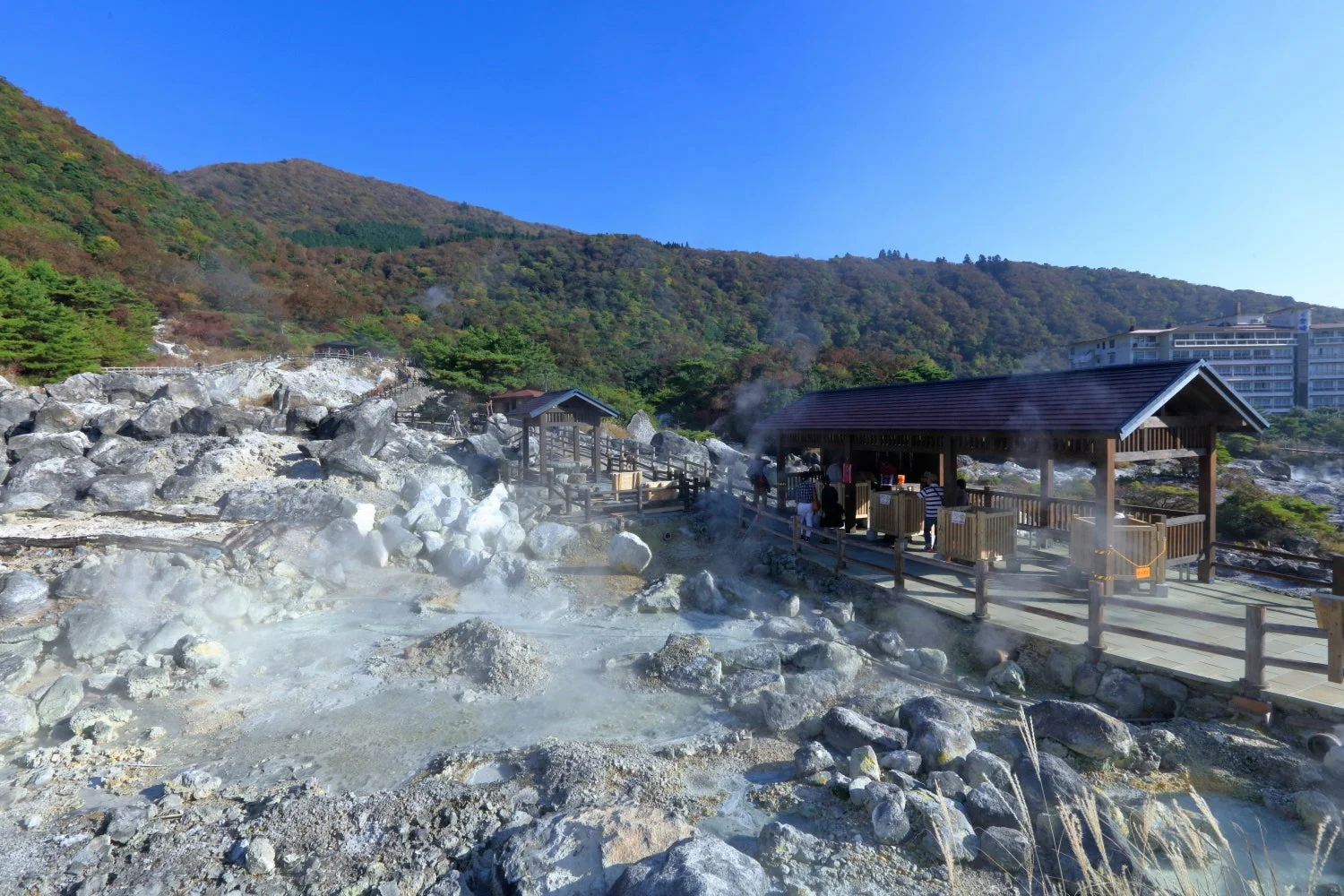 噴気が立ち上る火山性の景観が広がる温泉地です。周囲は紅葉が始まった山々に囲まれ、木製の散策路や休憩所が見え、遠方にはホテルらしき建物もあります。秋の穏やかな日差しの下、日本の自然豊かな観光地が描かれています。
