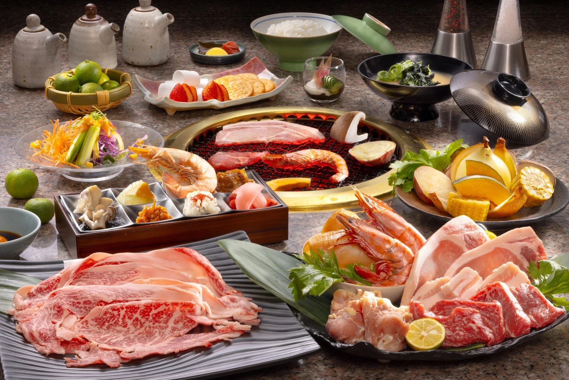 豪華な焼肉の食卓が写っており、牛肉、豚肉、鶏肉、海老、様々な野菜がグリルを囲んで並べられている