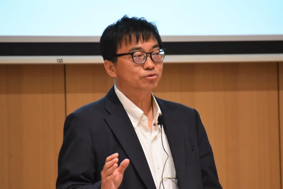 眼鏡をかけたアジア系の男性が、白いシャツとスーツを着用し、マイクを付けて講演またはプレゼンテーションを行っています。