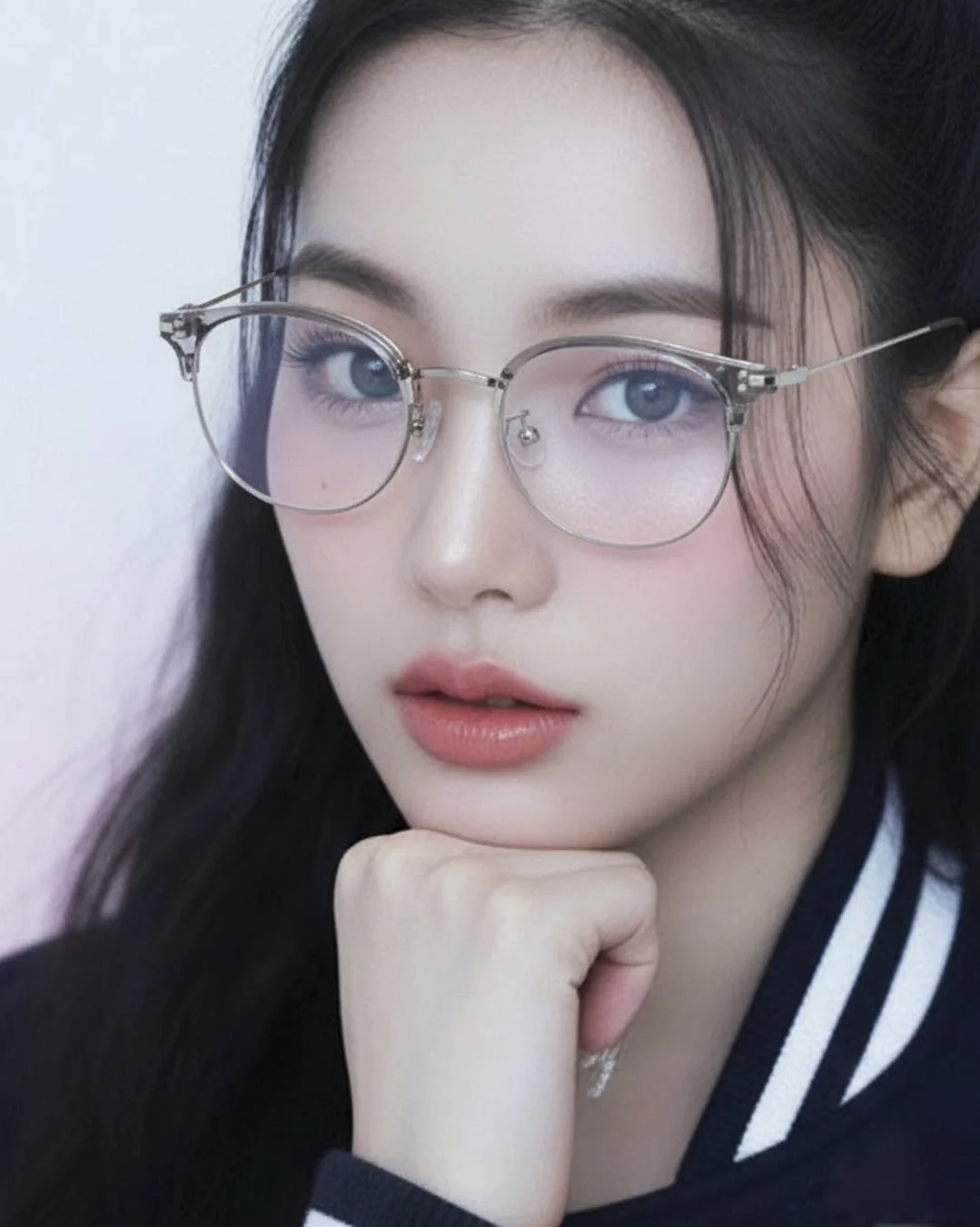 メガネをかけた若い女性のクローズアップポートレートです。彼女は青い瞳と長い黒髪を持ち、チークとリップメイクを施しています。顎に手を当てており、知的な雰囲気を醸し出しています。