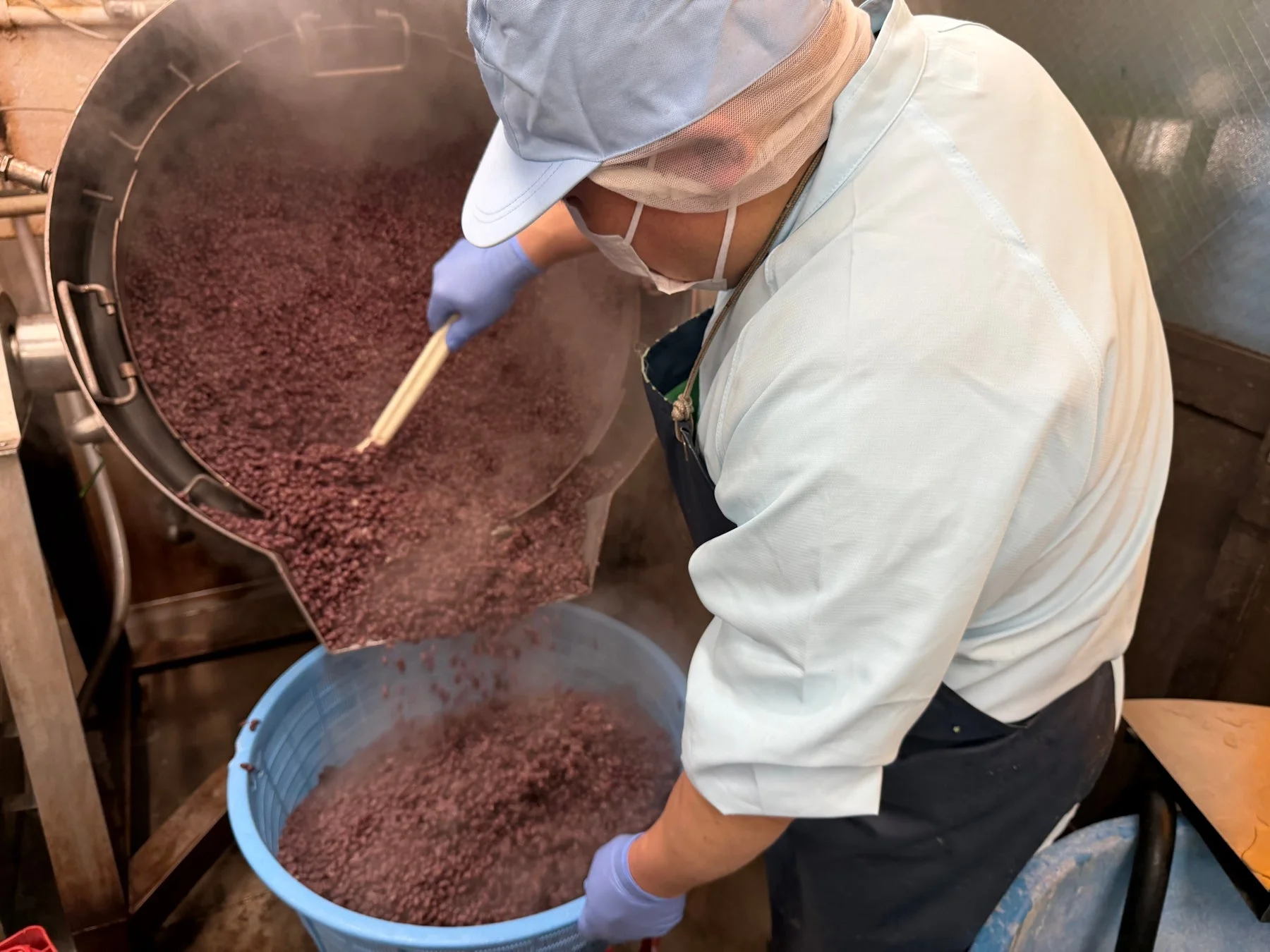 食品工場で、作業員が大きな機械から湯気の立つ小豆を青い容器に移している様子